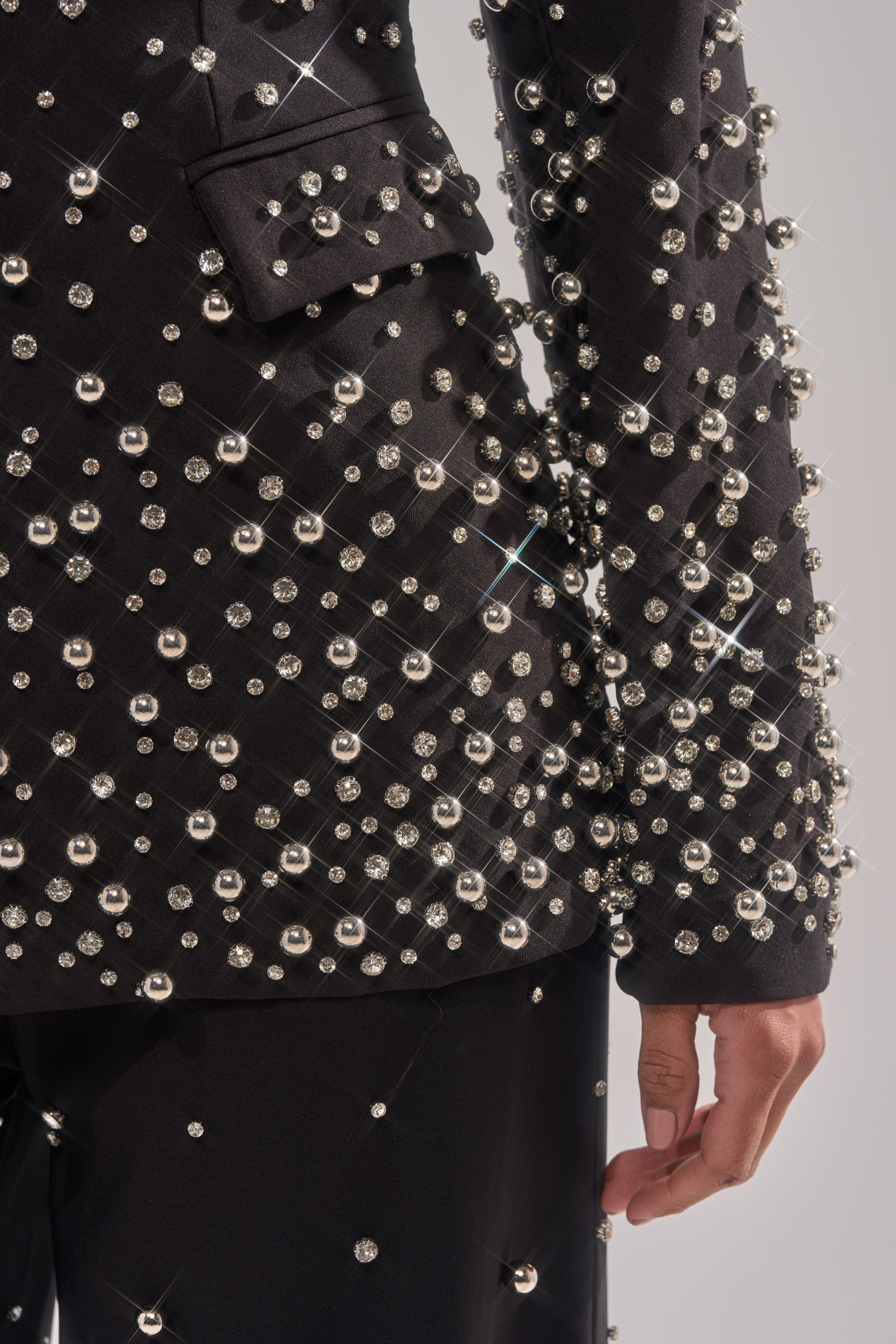 A close-up of a person wearing the GET CLOSER EMBELLISHED BLAZER, adorned with shiny silver studs that reflect light for a sparkling effect. Only the torso and one hand are visible.