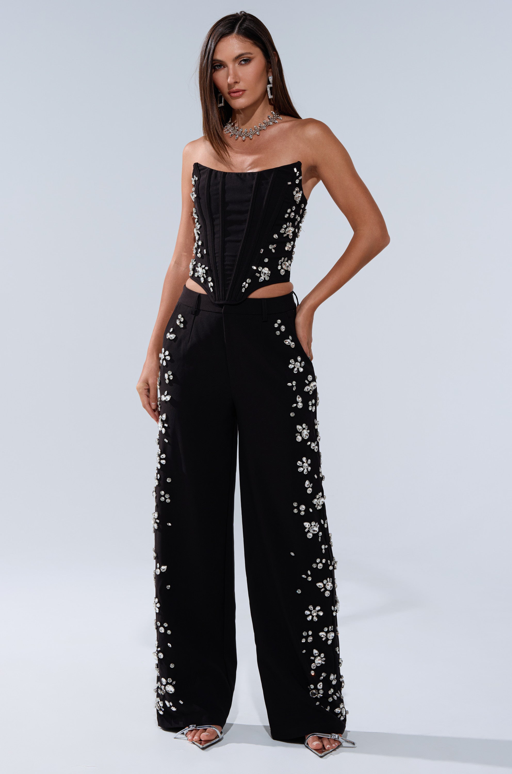 A woman poses in the FEELING RICH TROUSER WITH RHINESTONE, a strapless black jumpsuit with a corset-style bodice and high-rise wide-leg pants accented by white floral rhinestones, paired with silver heels and a statement necklace.