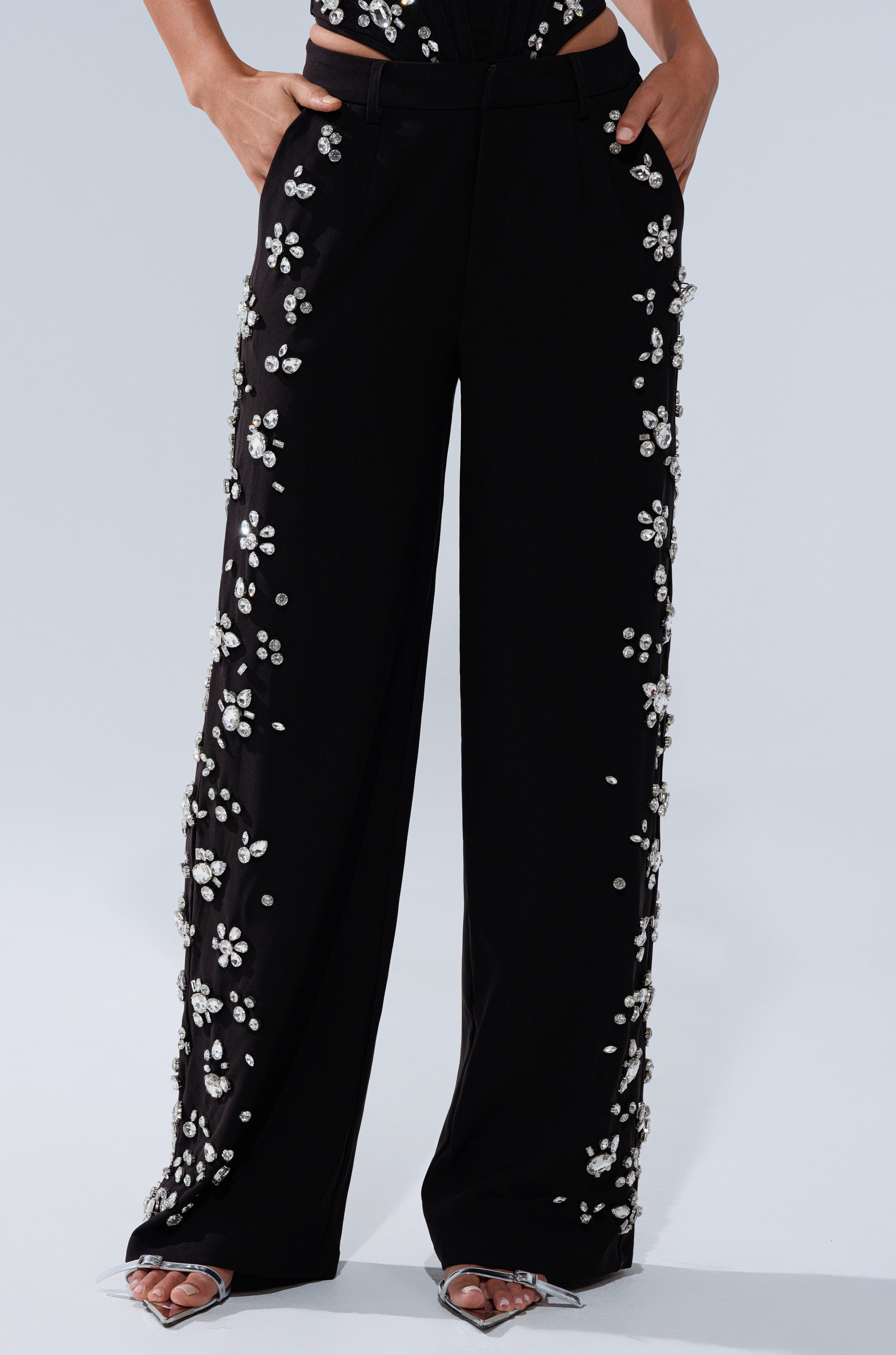 A woman stands in silver open-toe heels, hands in her pockets, wearing FEELING RICH TROUSER WITH RHINESTONE—high-rise, wide-leg pants featuring white floral rhinestone embellishments.