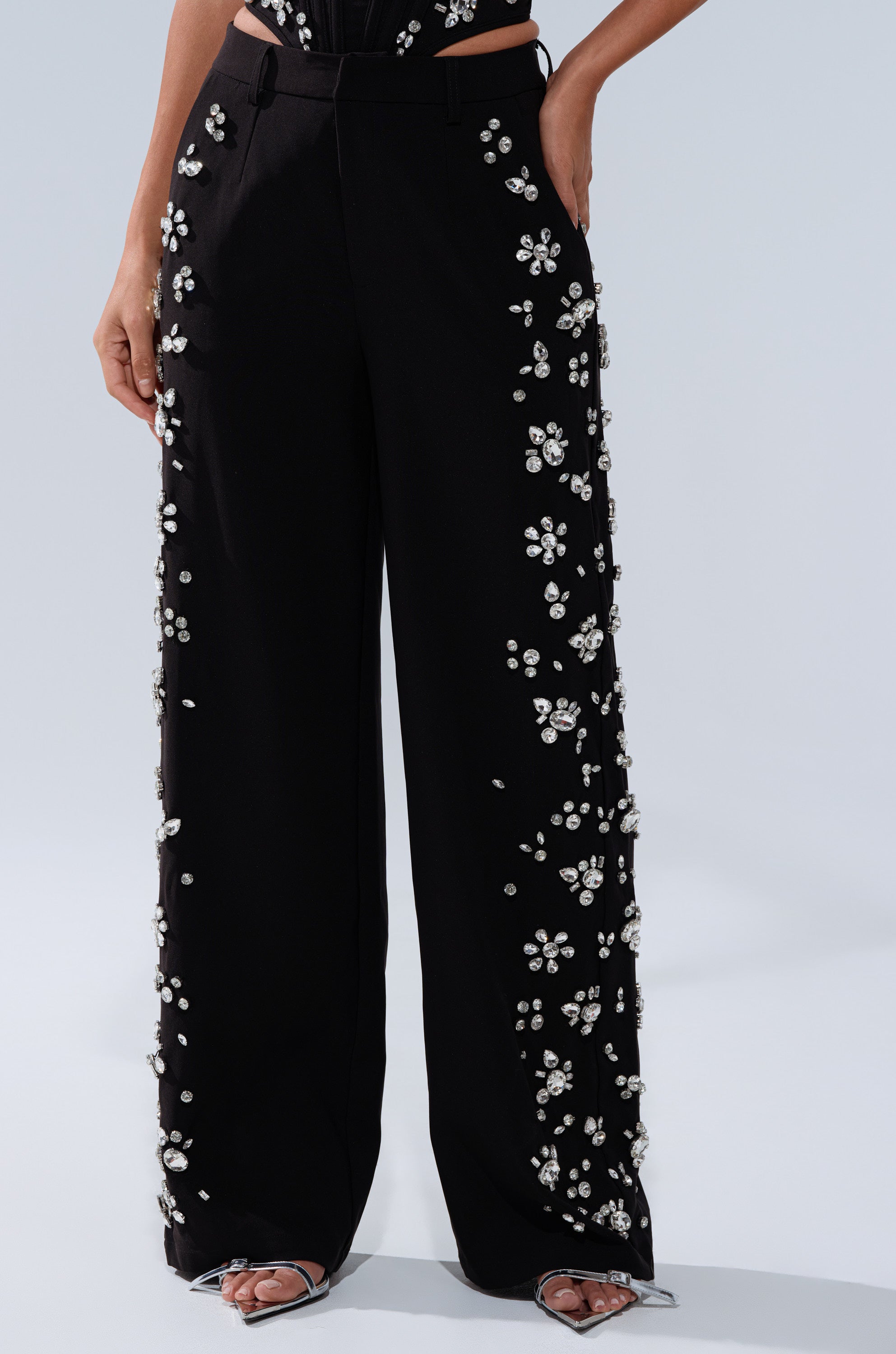 A person wears black FEELING RICH TROUSER WITH RHINESTONE—high-rise wide-leg pants embellished with sparkling rhinestones along the sides and front—paired with open-toed silver heels.