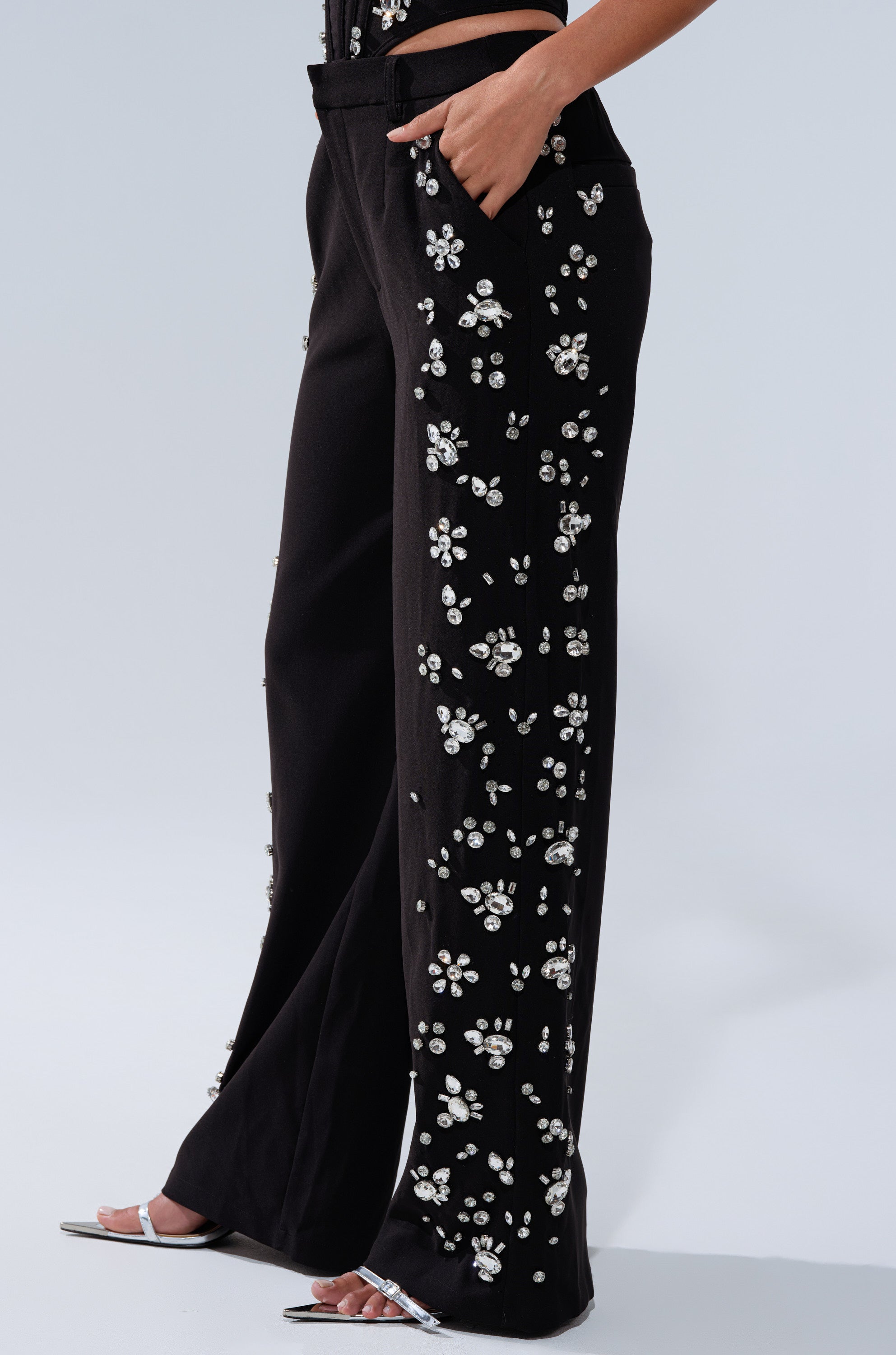 A person models the FEELING RICH TROUSER WITH RHINESTONE—black trousers adorned with white floral rhinestones—paired with silver open-toe heels, posing confidently with one hand in the pocket.