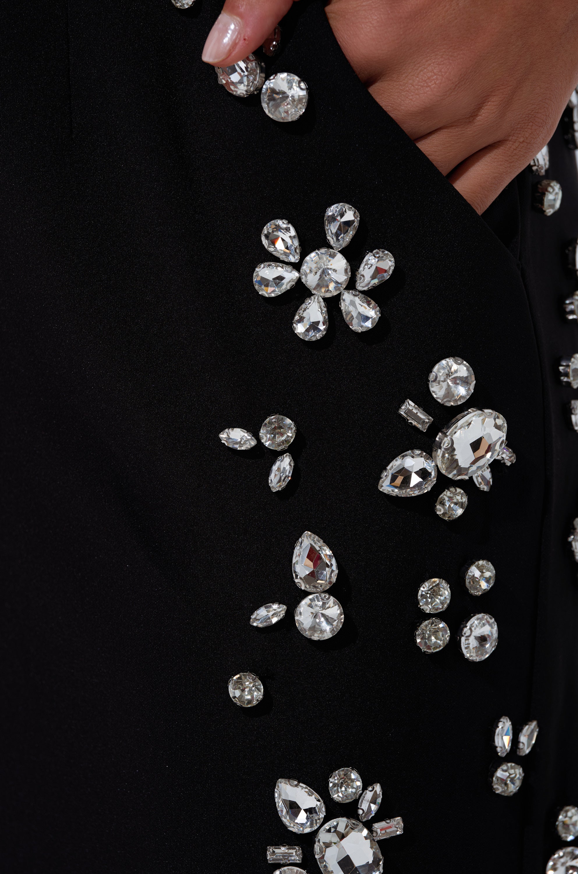 A close-up of manicured nails resting in the pocket of sleek black FEELING RICH TROUSER WITH RHINESTONE—high-rise wide-leg pants embellished with clusters of large, shiny rhinestones in elegant floral patterns.