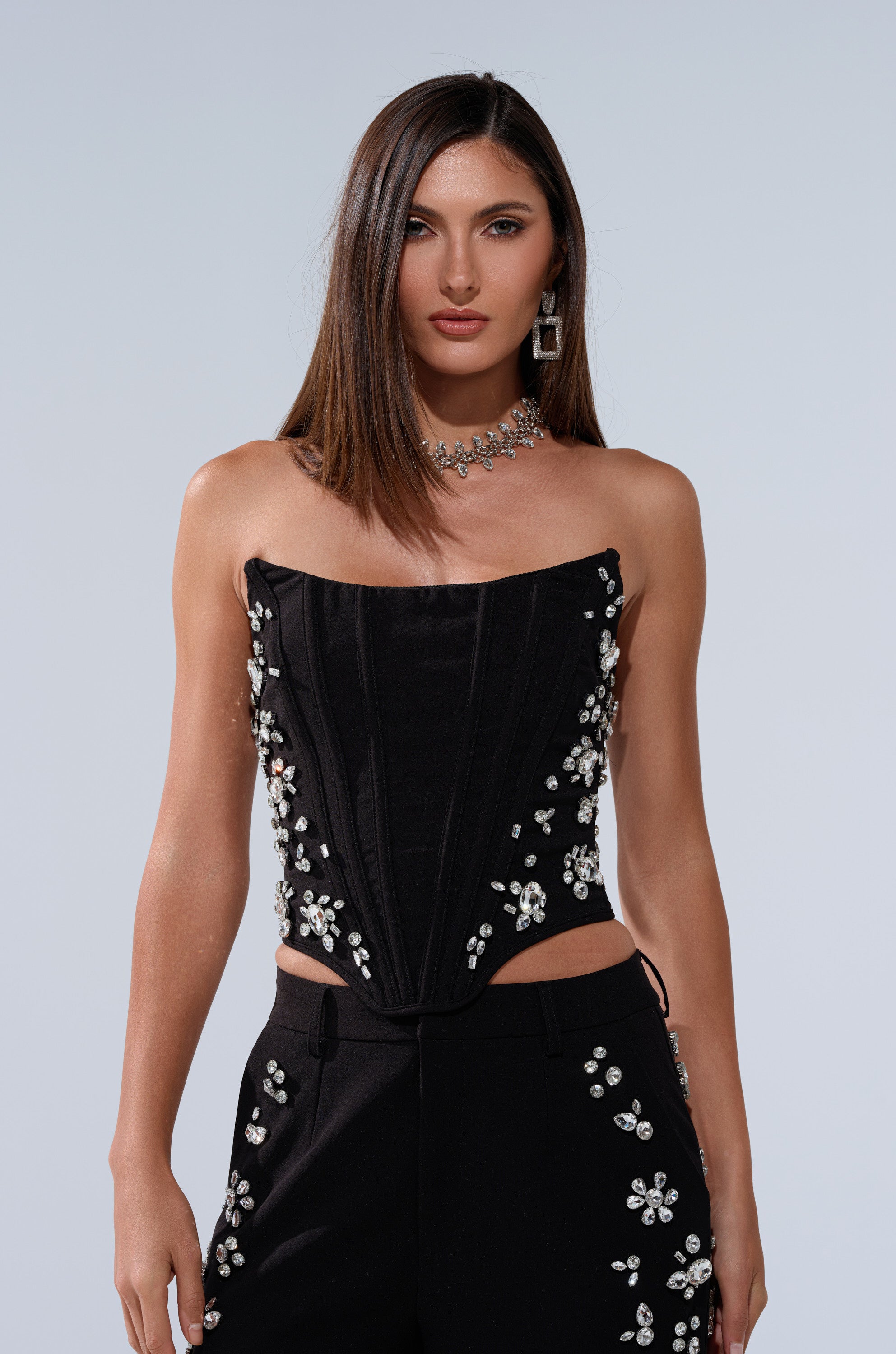 A woman with straight brown hair wears the FEELING RICH CORSET WITH RHINESTONE, a strapless black top featuring shiny floral embellishments. The curve-hugging fit highlights her silhouette as she accessorizes with jeweled necklace and earrings. 2