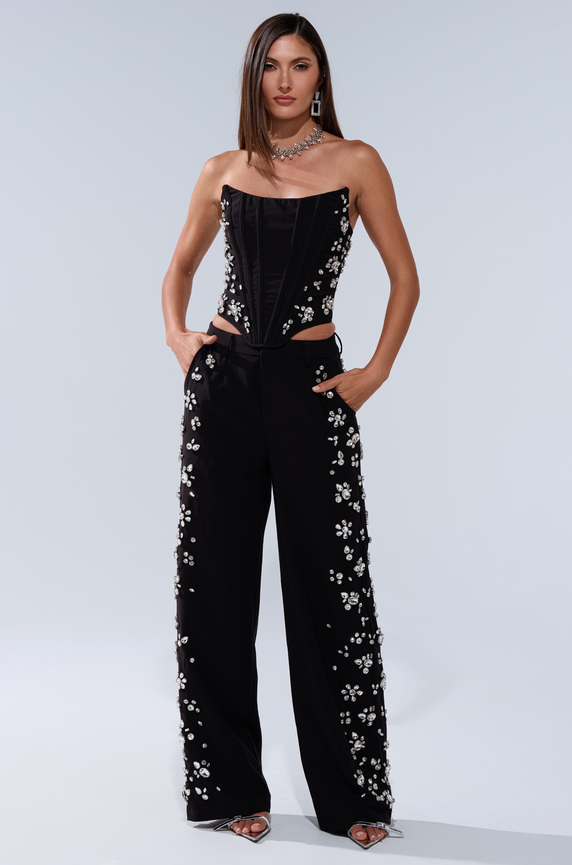 A woman exudes confidence in the FEELING RICH CORSET WITH RHINESTONE, a strapless black top with a sleek fit, styled with high-waisted black pants featuring white floral accents, straight hair, a statement necklace, and open-toe heels. 6