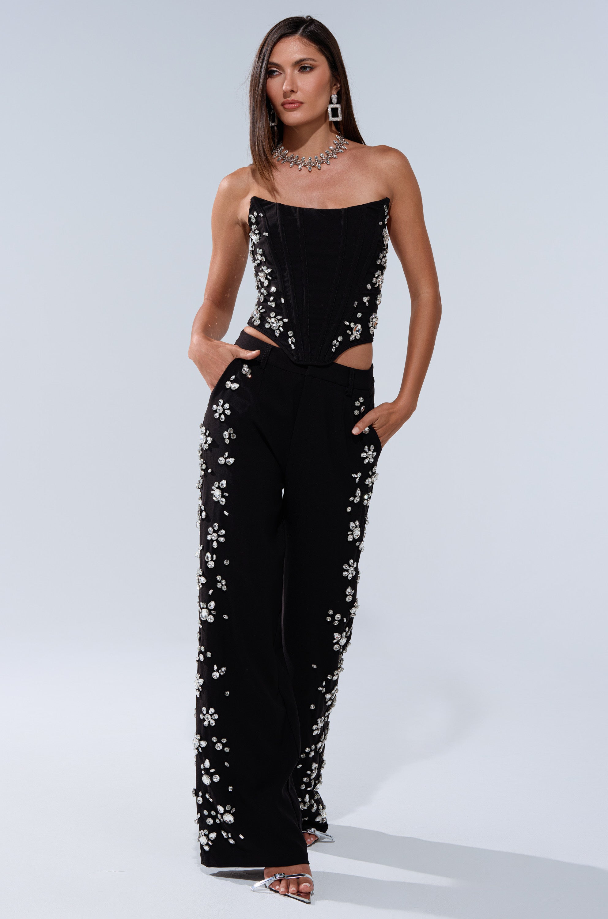 A woman wears the FEELING RICH CORSET WITH RHINESTONE, a strapless black jumpsuit with a curve-hugging fit, wide-leg pants, and rhinestone embellishments. She pairs it with statement jewelry and silver heels against a plain light background. 7