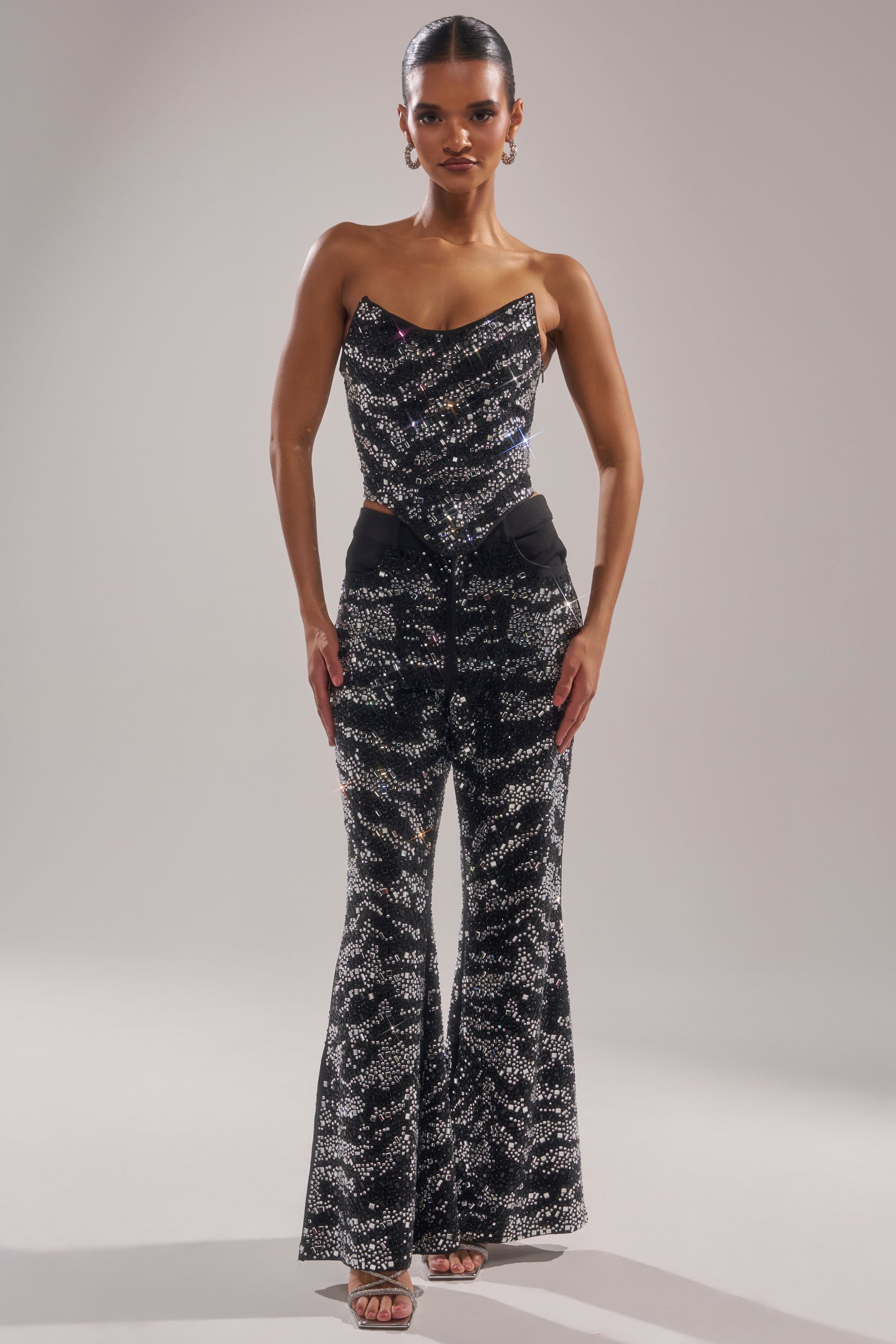 A woman stands confidently in the TIME TO CELEBRATE AND DANCE RHINESTONE TROUSER—a high-rise, wide-leg pant featuring a shimmering abstract pattern—paired with a matching black sequined strapless top, set against a plain light background.