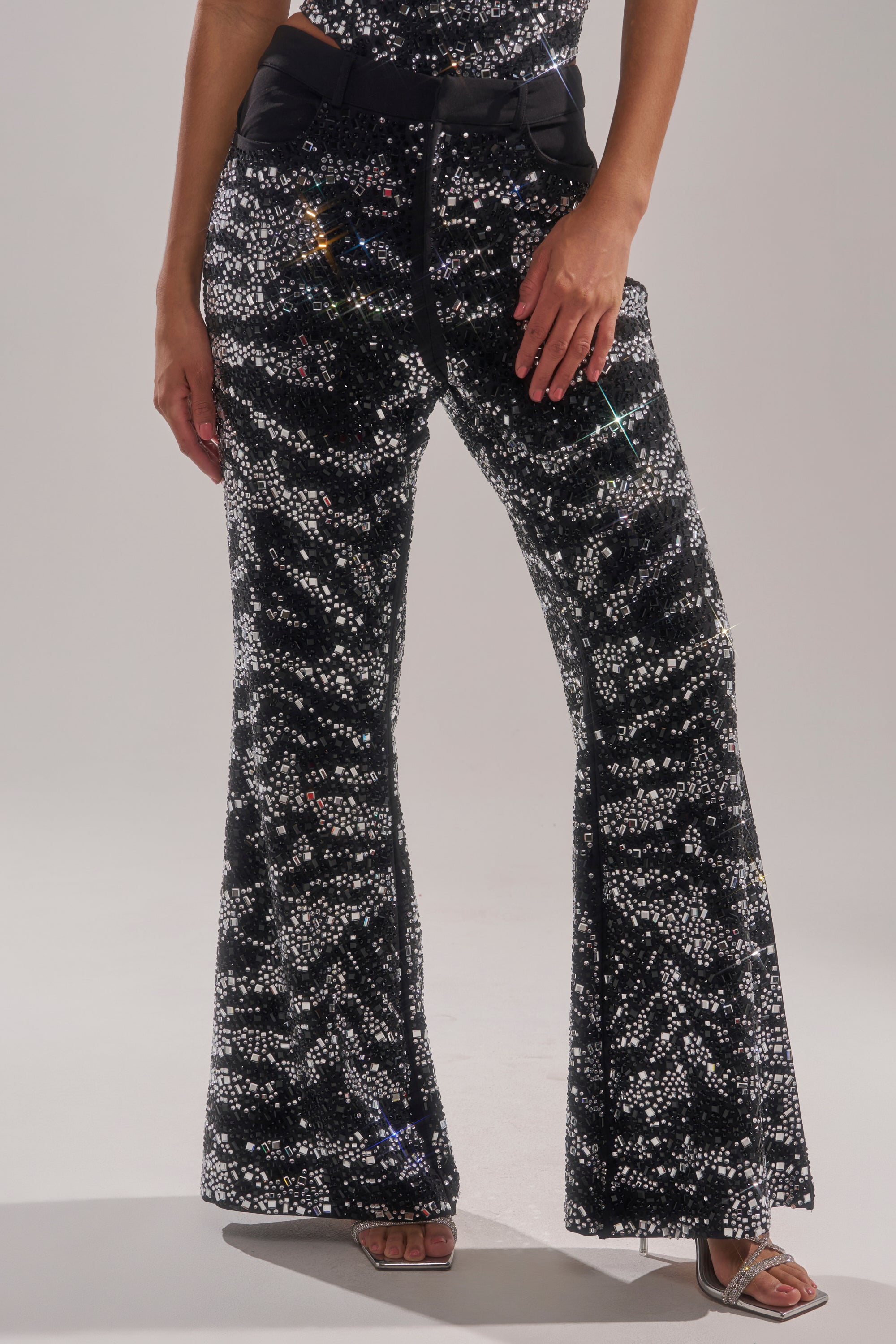 A person wearing the TIME TO CELEBRATE AND DANCE RHINESTONE TROUSER—high-waisted black sequin flared pants with a silver and black pattern—paired with heeled sandals, stands against a plain light background.
