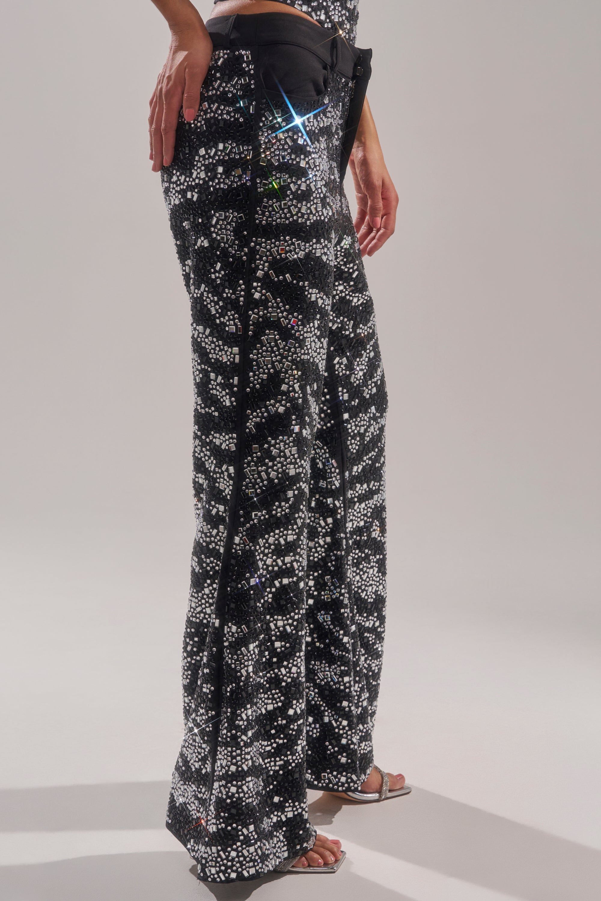 A person wears the TIME TO CELEBRATE AND DANCE RHINESTONE TROUSER—high-rise, black and white sequin pants with a pixelated pattern and flared legs—paired with open-toed heels in a neutral studio, making the sparkling trousers stand out.