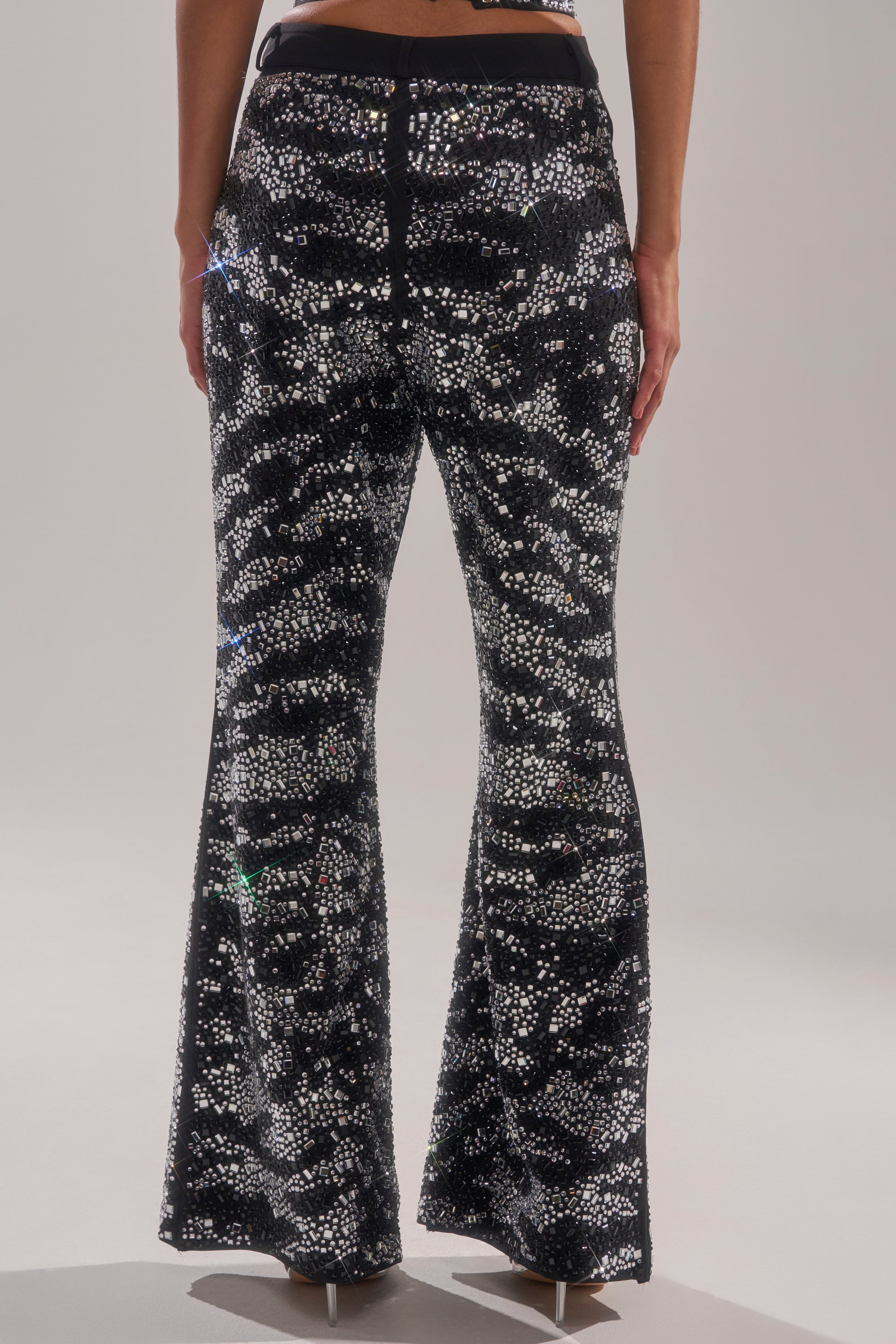 Person models the TIME TO CELEBRATE AND DANCE RHINESTONE TROUSER, featuring a high-rise fit and black flared-leg silhouette adorned with black and silver sequins in a sparkly, camouflage-like pattern; image shows the trousers from the back.