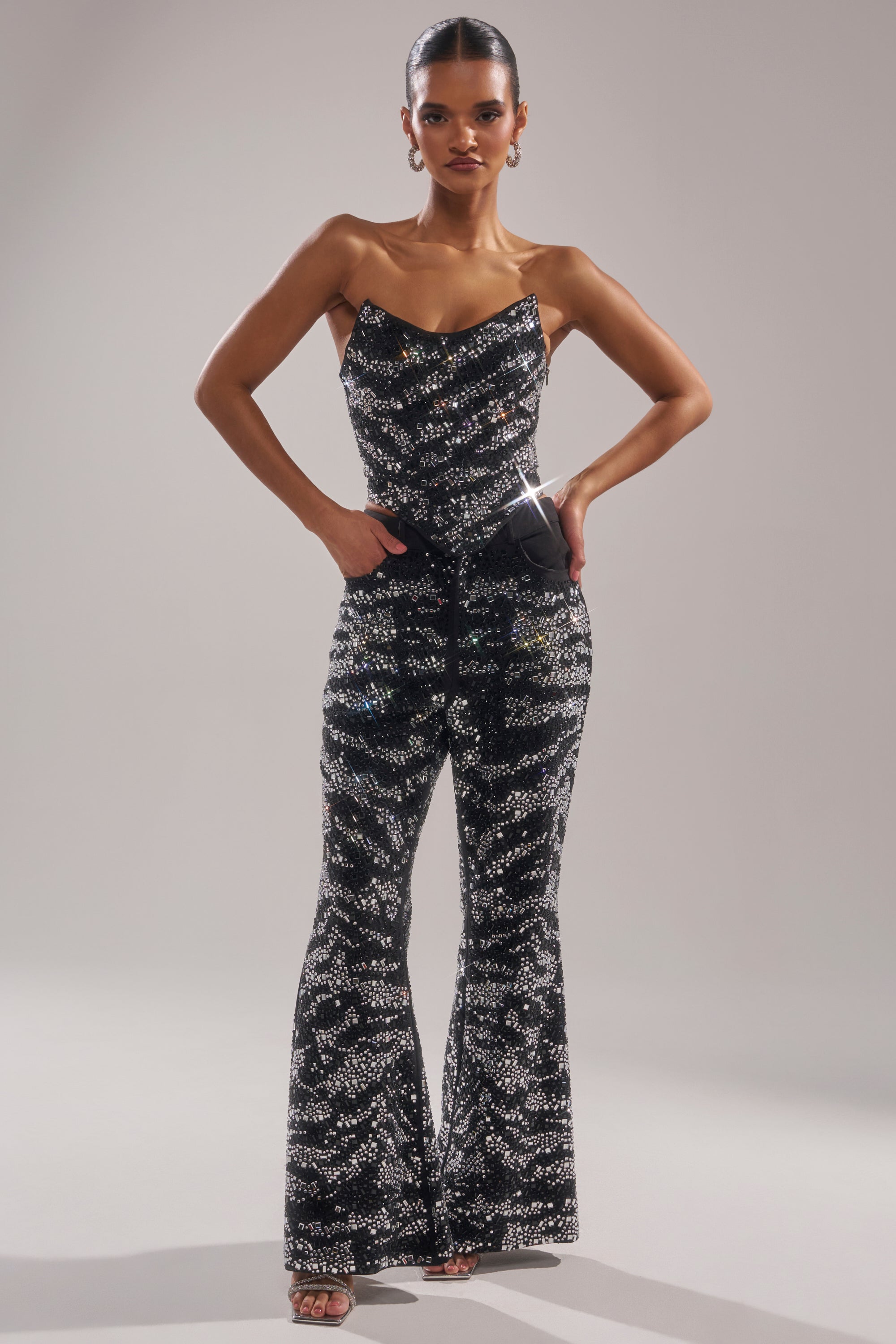 A woman exudes confidence in the TIME TO CELEBRATE AND DANCE RHINESTONE TROUSER, featuring a black, sequined finish and flared legs. With slicked-back hair and a chic black clutch, she completes her striking look.