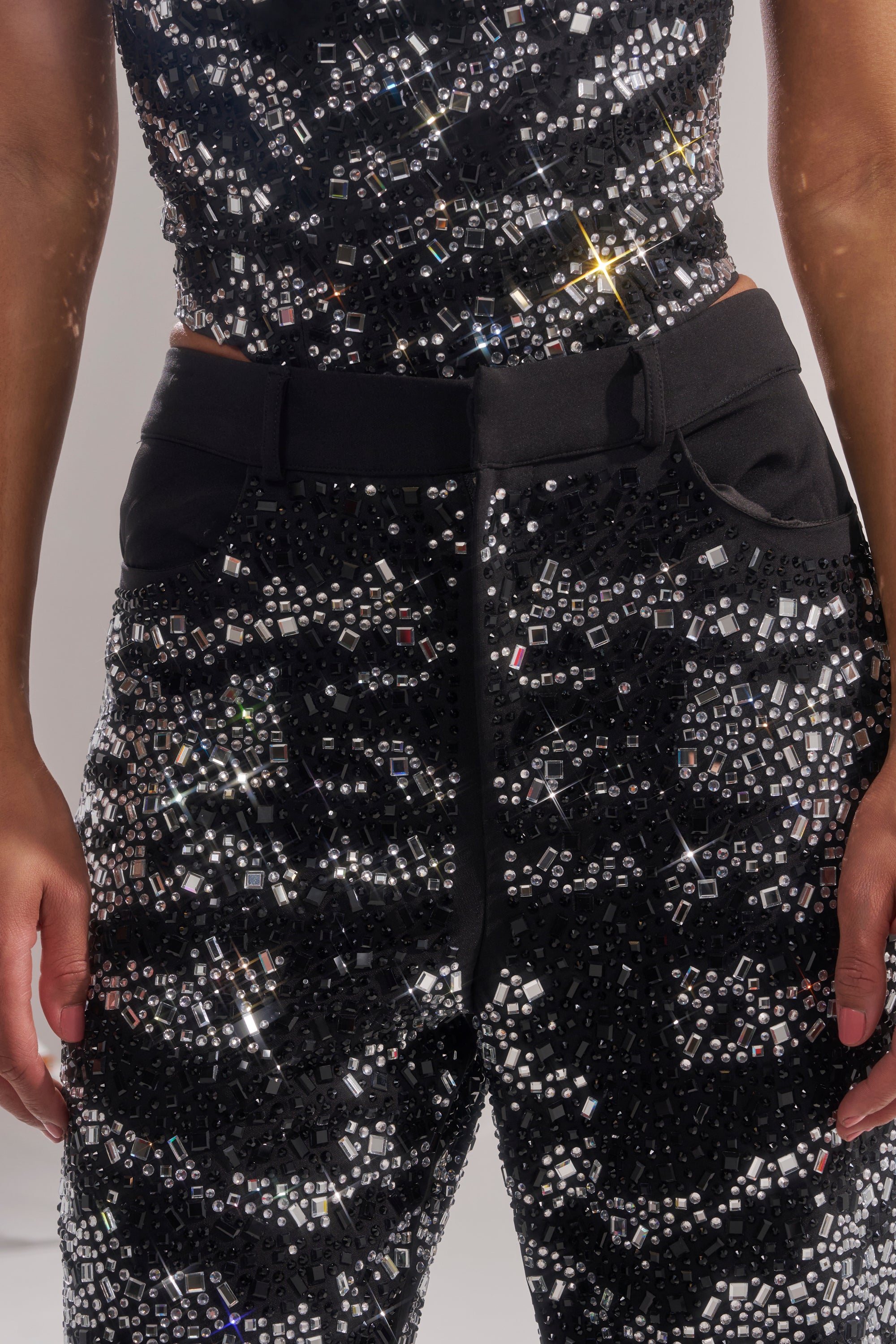 A person wears the TIME TO CELEBRATE AND DANCE RHINESTONE TROUSER, a sparkling black high-rise fit covered in shiny sequins and rhinestones, with arms partially visible and relaxed hands at their sides.
