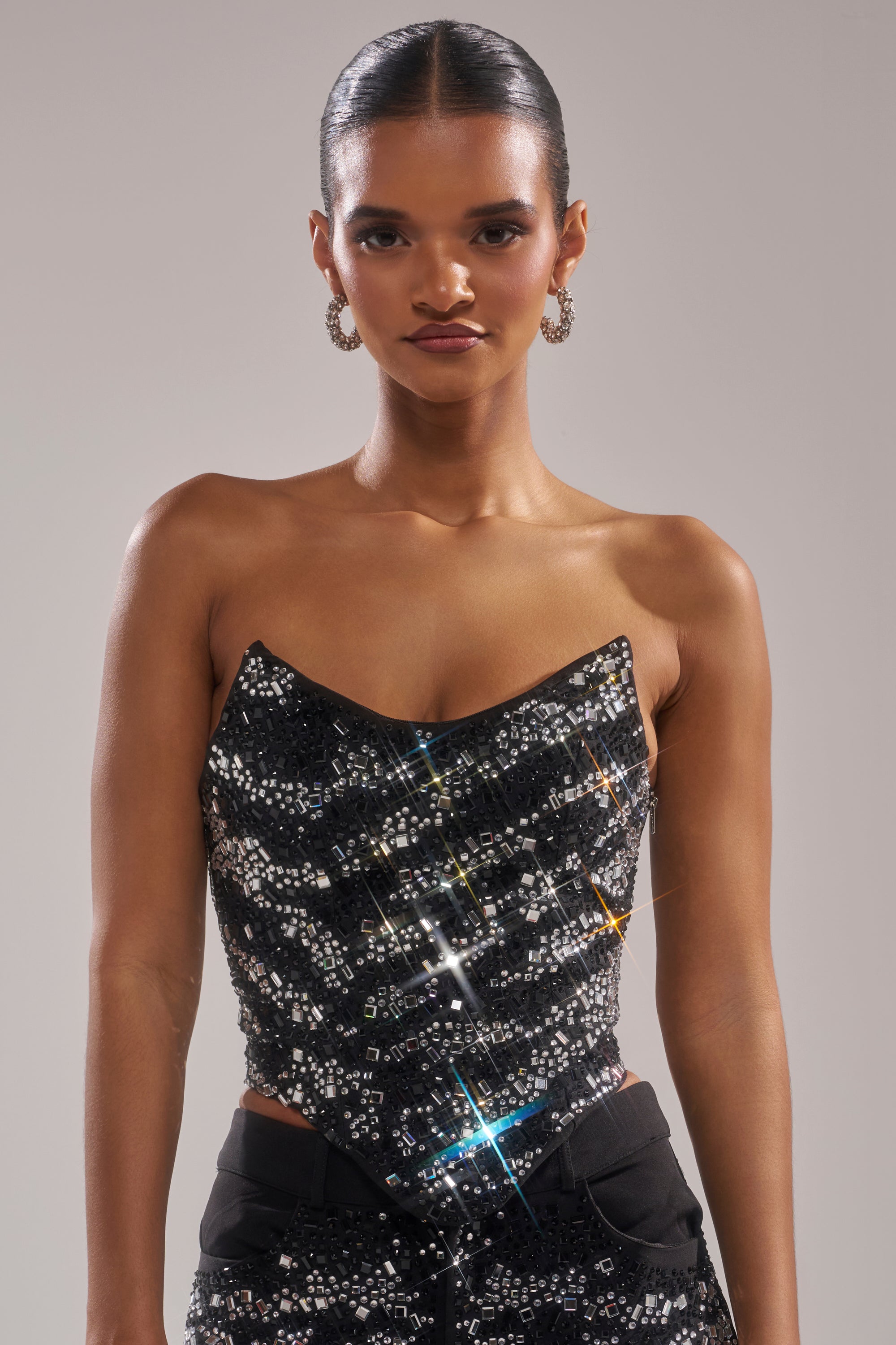 A woman with slicked-back hair wears the TIME TO CELEBRATE AND DANCE RHINESTONE CORSET and matching sequin pants. She accessorizes with hoop earrings and poses confidently against a plain light gray background.