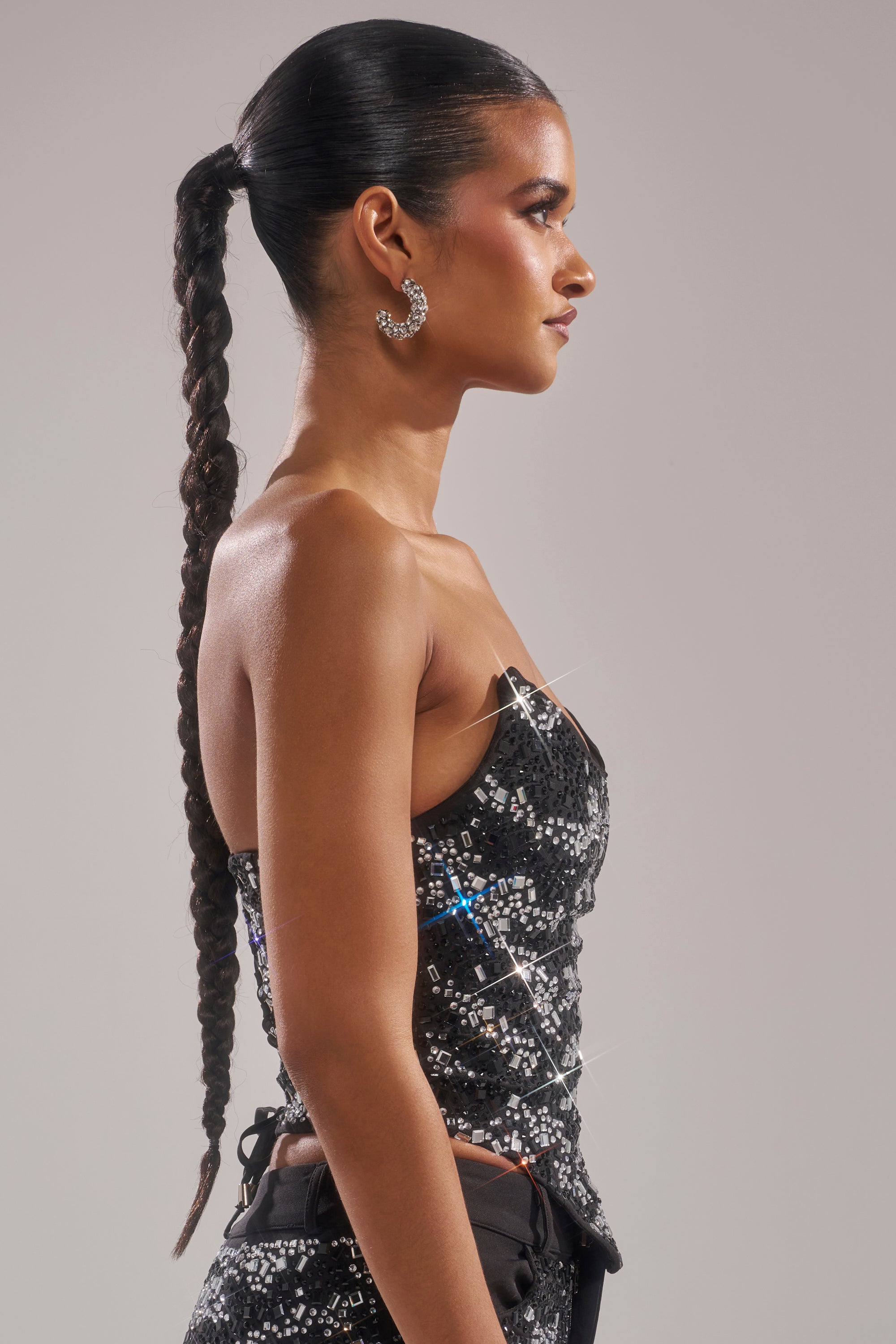 Wearing the TIME TO CELEBRATE AND DANCE RHINESTONE CORSET, a woman with a long braided ponytail and sparkly hoop earrings stands in profile against a plain background.