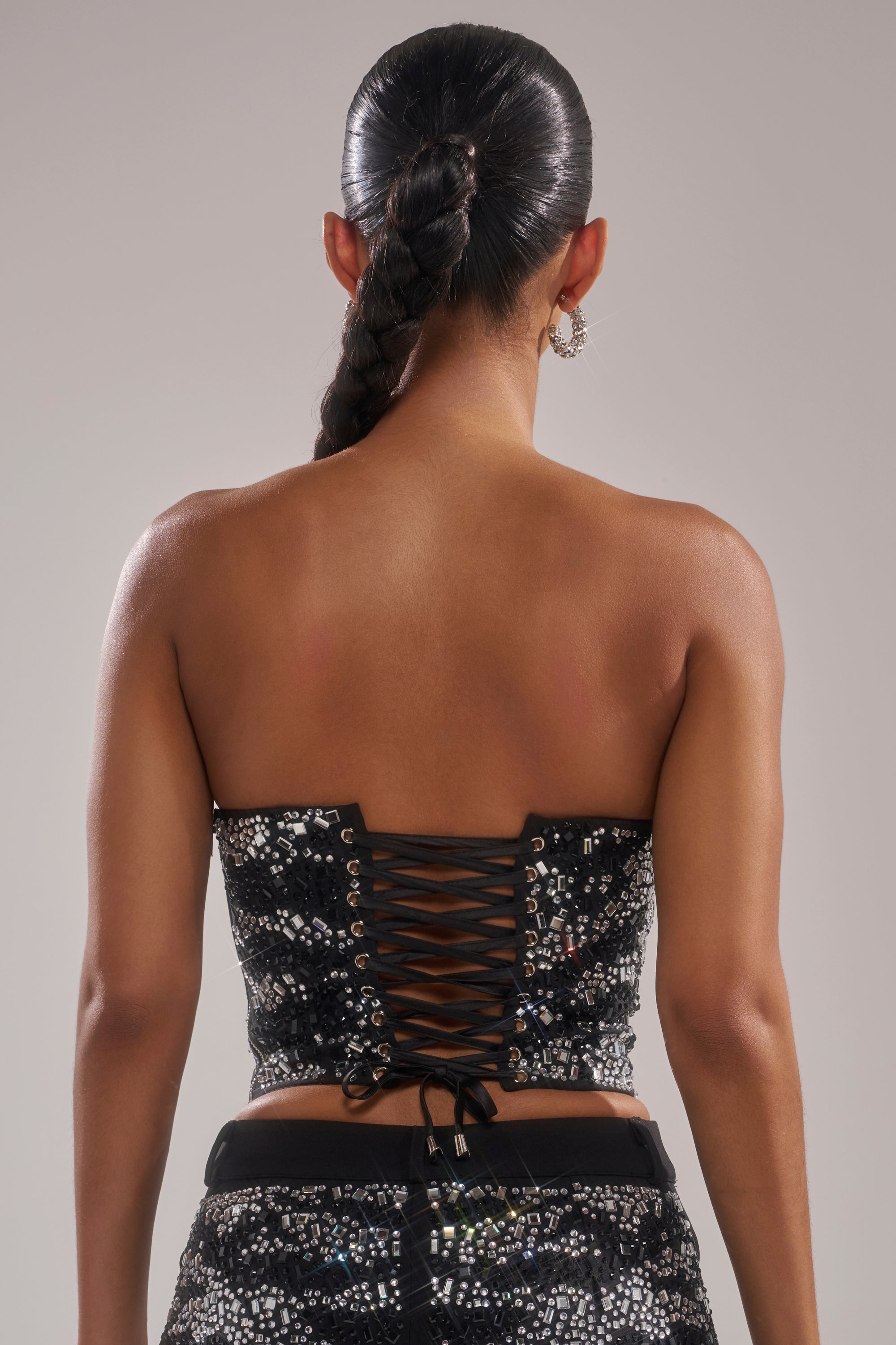 A woman with a long braided ponytail is seen from the back, wearing the TIME TO CELEBRATE AND DANCE RHINESTONE CORSET and matching sequined pants. She accessorizes with hoop earrings and poses against a neutral background.