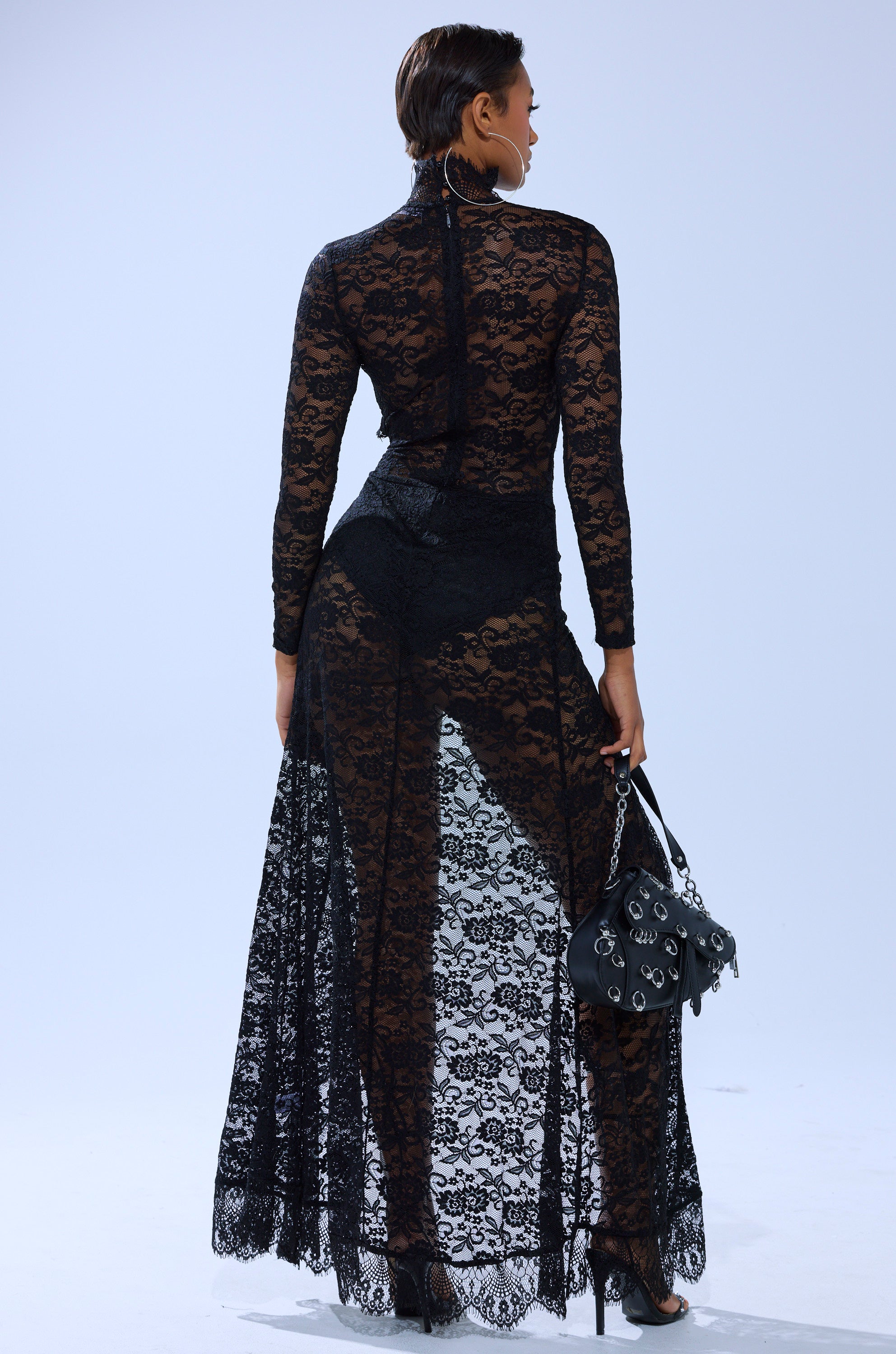 A woman faces away in the LAY IT LOW LACE MAXI DRESS, featuring sheer floral lace over black undergarments. She accessorizes with a black purse adorned with silver charms, black heels, and neat short hair.