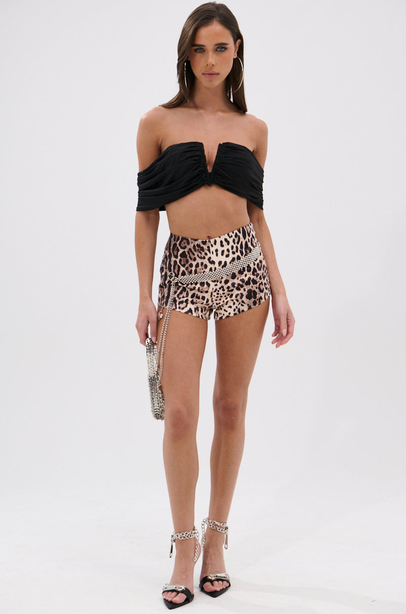 A woman stands against a plain white background wearing the LOVING YOU IS EASY CHEETAH SHORTS, a black off-shoulder crop top, silver high-heeled sandals, hoop earrings, and holding a small silver purse.