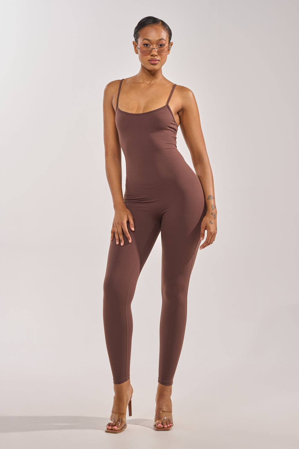 A woman with short hair and sunglasses poses confidently against a plain light background, wearing the THAT GIRL SEAMLESS JUMPSUIT IN BROWN with a skinny-leg silhouette and transparent high-heeled sandals. 1