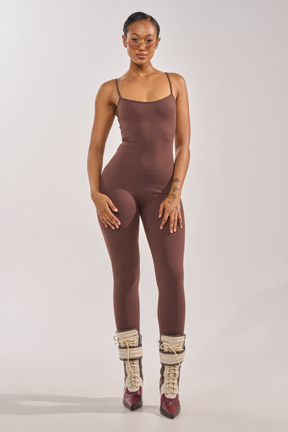 A person stands facing forward, wearing the THAT GIRL SEAMLESS JUMPSUIT IN BROWN, brown and cream lace-up boots, clear glasses, and earrings, posing against a plain light background. 3