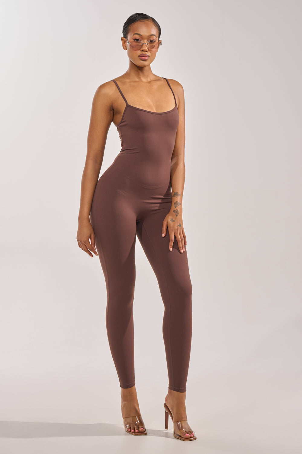 A woman poses against a plain light background in the THAT GIRL SEAMLESS JUMPSUIT IN BROWN, styled with clear heels and light pink sunglasses, showcasing the chic appeal of this fitted, sleeveless jumpsuit with spaghetti straps. 4