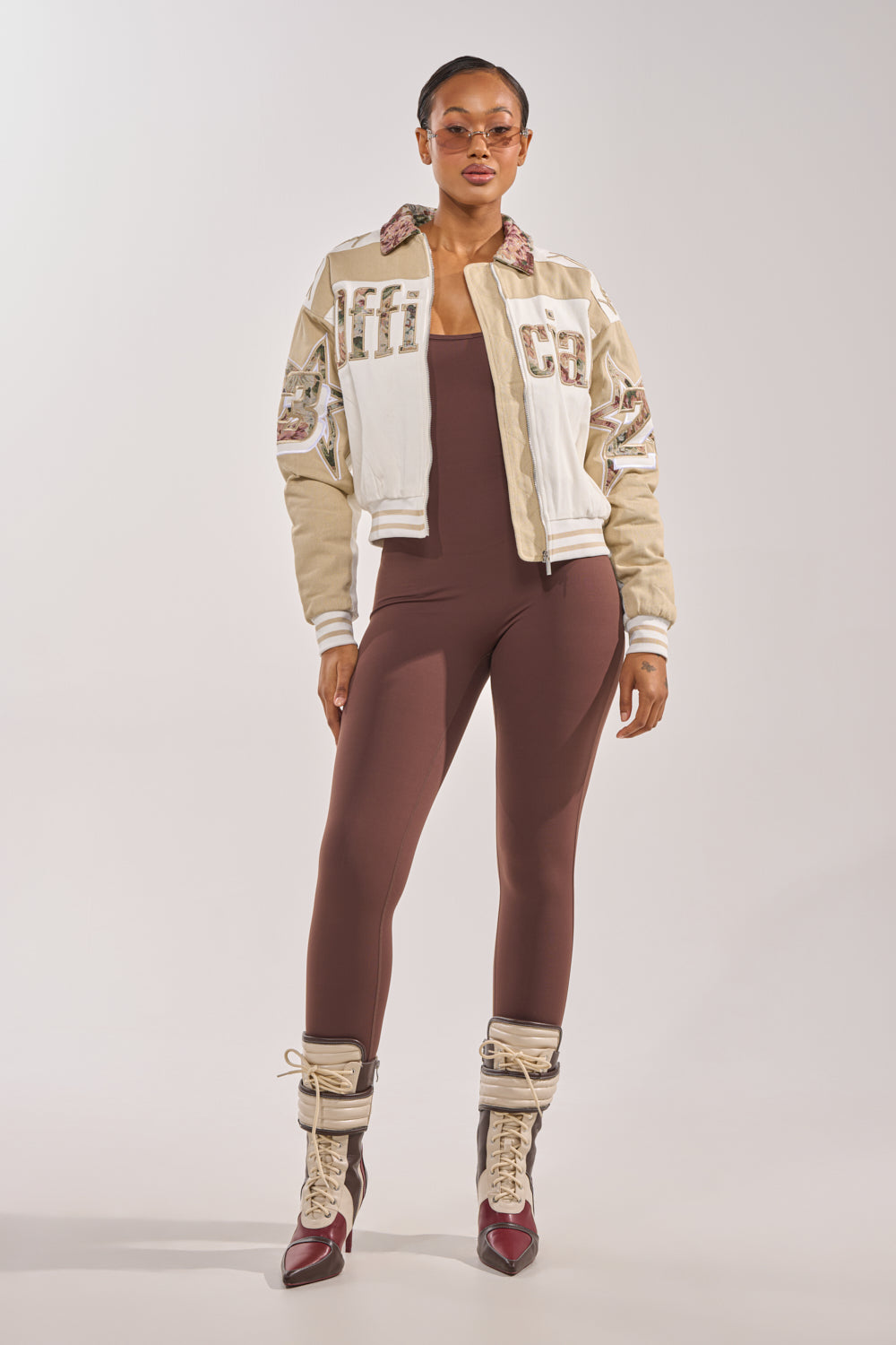 A woman stands confidently, facing forward against a plain light background, wearing the THAT GIRL SEAMLESS JUMPSUIT IN BROWN styled with sunglasses, a cream and beige varsity jacket, and lace-up heeled boots. 5