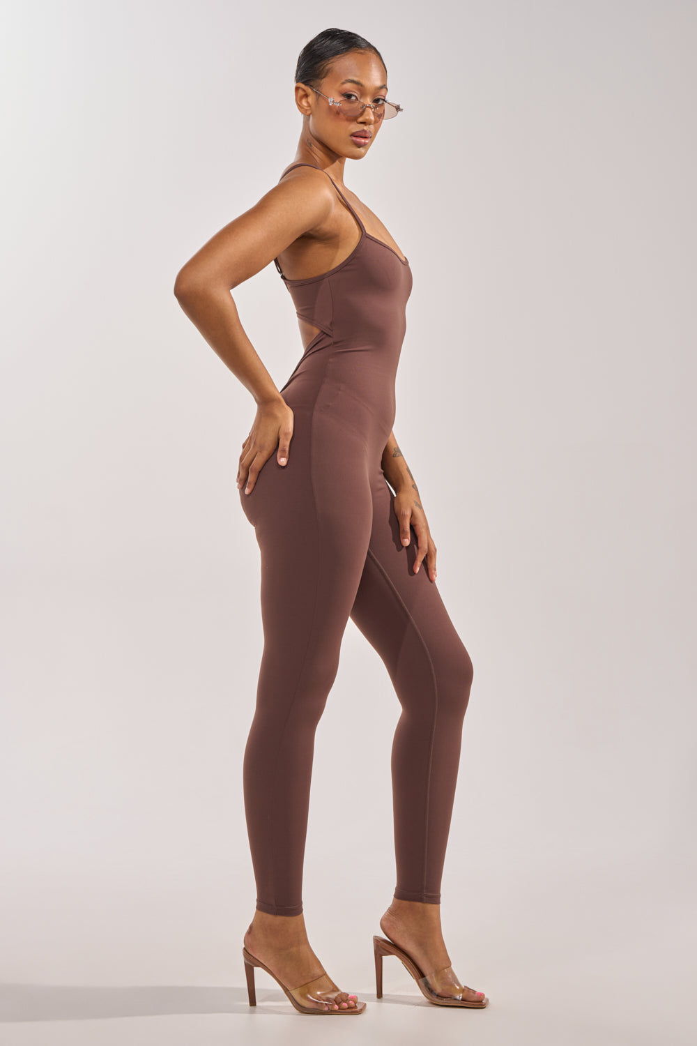 A woman models the THAT GIRL SEAMLESS JUMPSUIT IN BROWN, posing in profile with one hand on her hip. She completes the look with clear heels and round glasses against a plain, light background. 6
