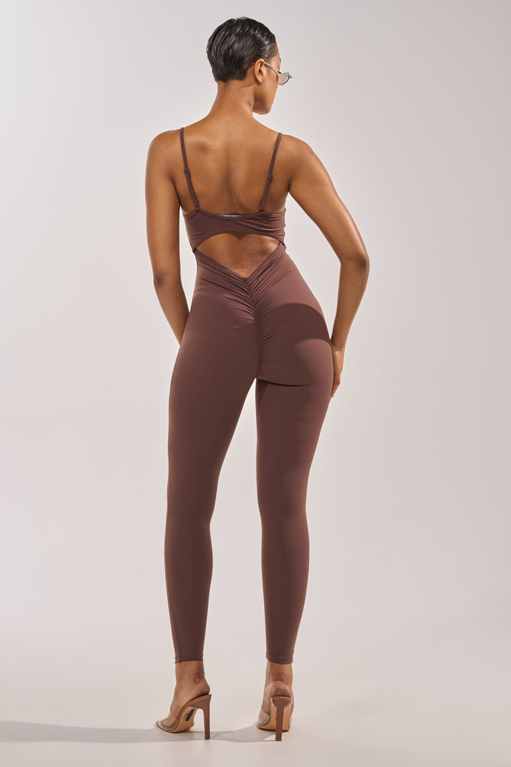 A person stands on high heels facing away, wearing the THAT GIRL SEAMLESS JUMPSUIT IN BROWN, featuring thin straps, an open-back cutout, and gathered seam detailing against a plain light gray background. 7
