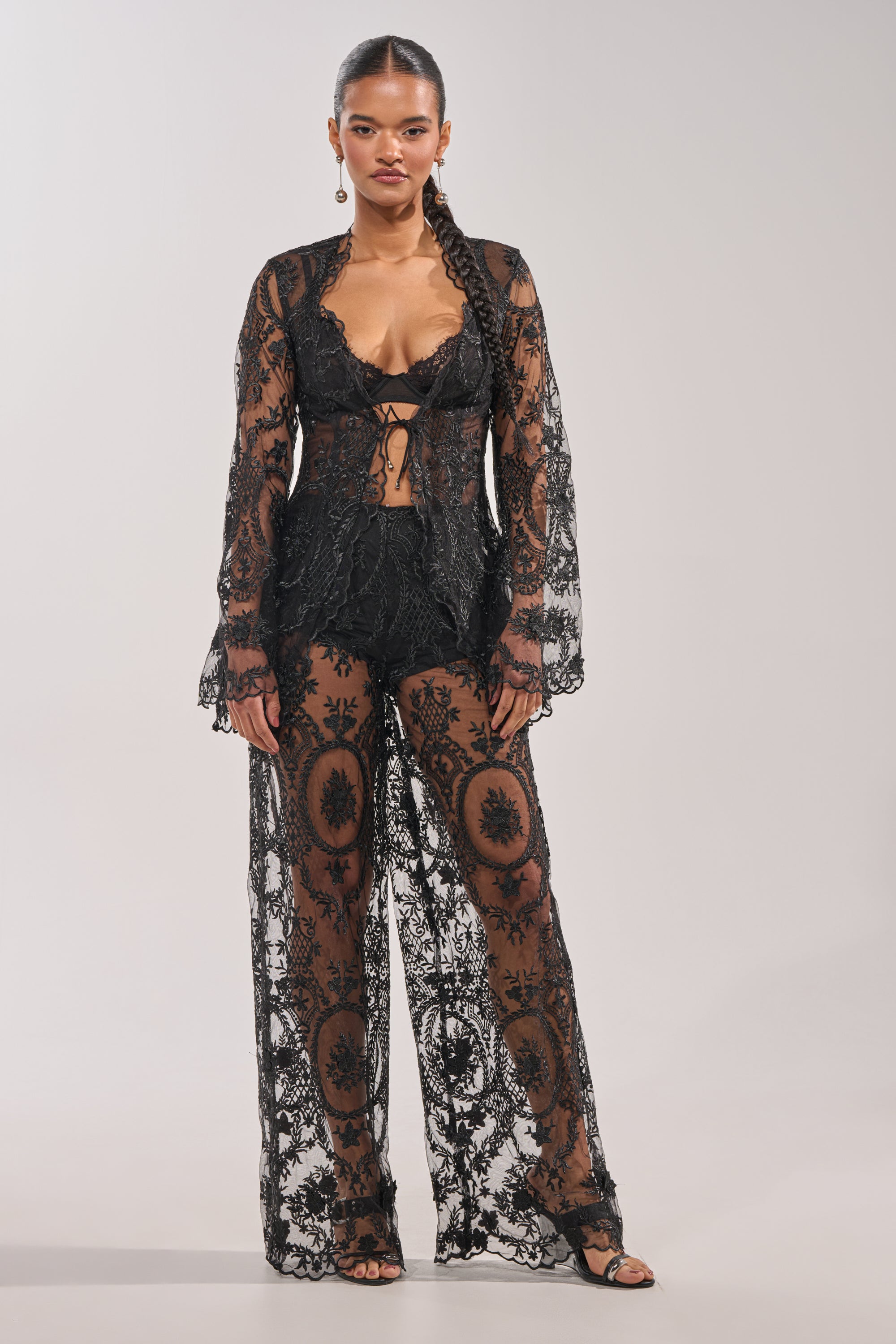 A woman with a long braid and confident expression models the SUPER LUXE WIDE-LEG PANT in sheer black lace with intricate patterns, paired with a matching long-sleeve bralette, styled for evening elegance. 7