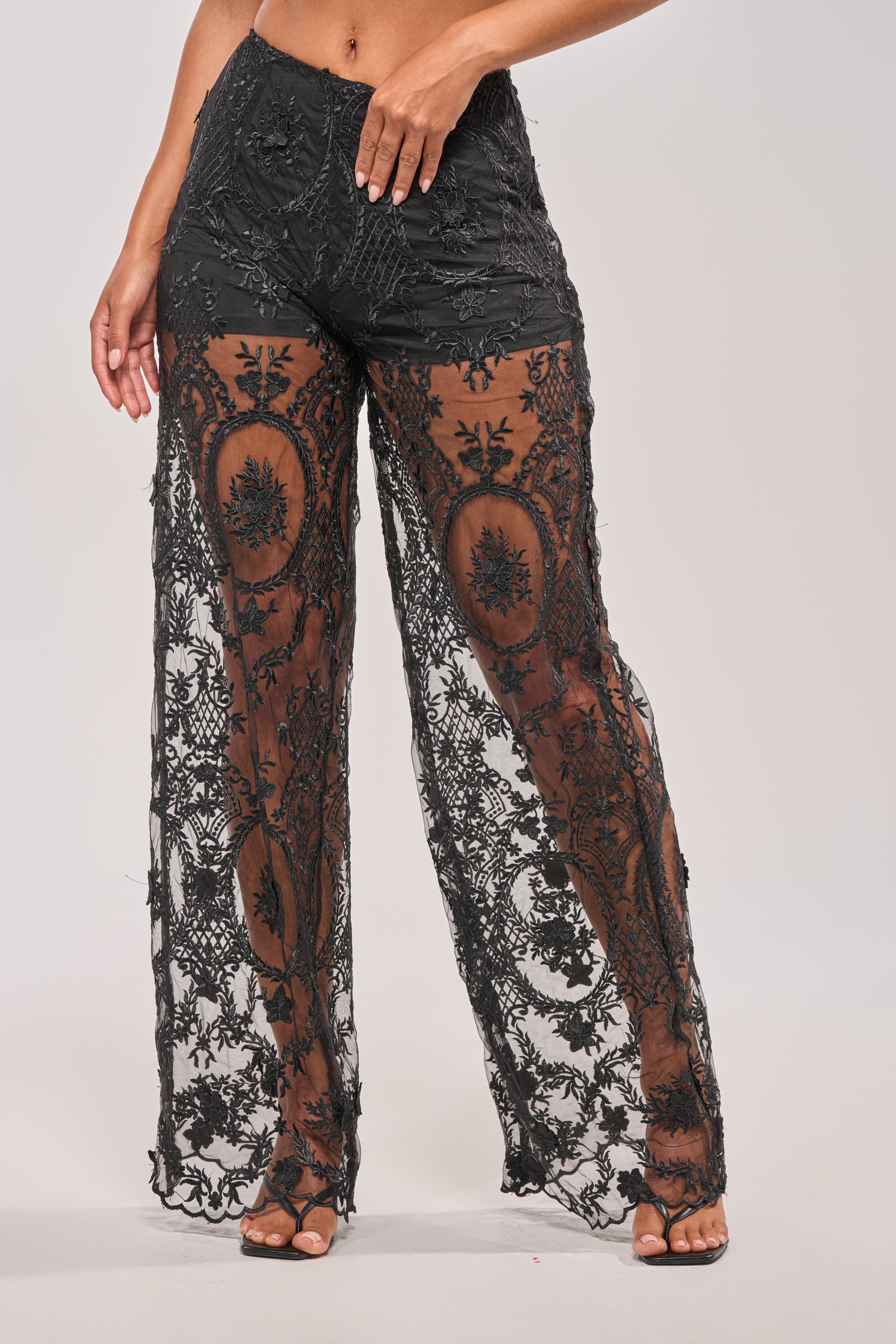 A person wearing SUPER LUXE WIDE-LEG PANT—black sheer lace pants with intricate floral embroidery over black shorts—stands barefoot, one hand on their waist. 1