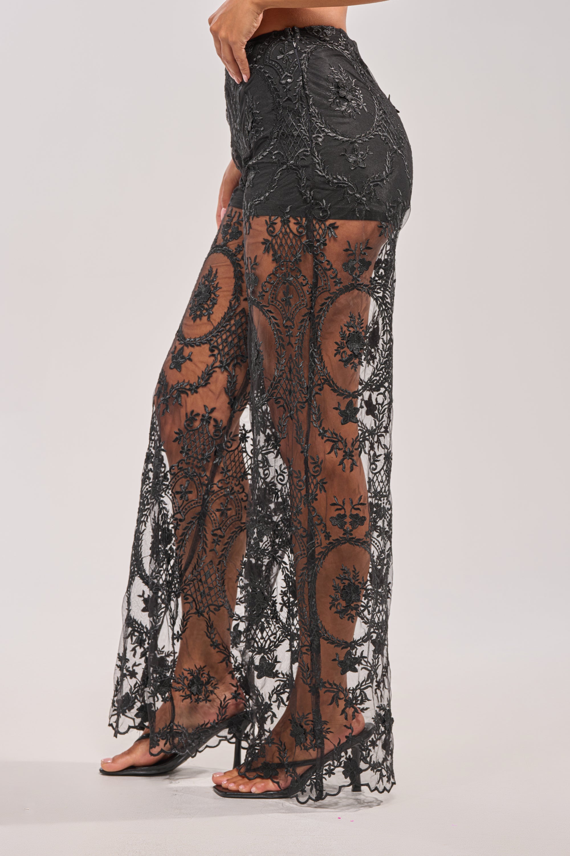 Model wears SUPER LUXE WIDE-LEG PANT, a high-rise black sheer lace pant with intricate floral patterns and black underwear underneath, paired with black sandals. Background is plain and light-colored. 3