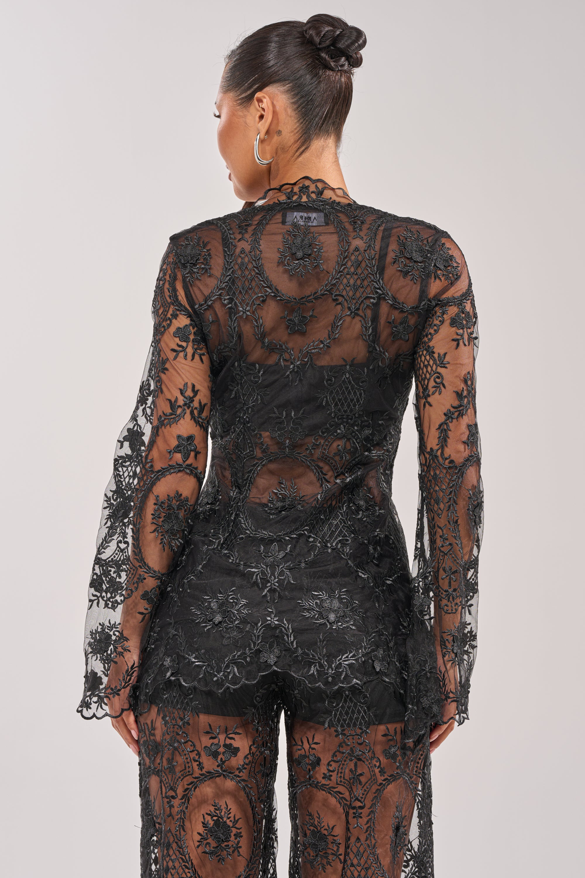 A woman with dark hair in a bun wears the SUPER LUXE LACE TIE-FRONT BLOUSE—a black sheer top with intricate floral and circular patterns—seen from the back against a plain light background. 4