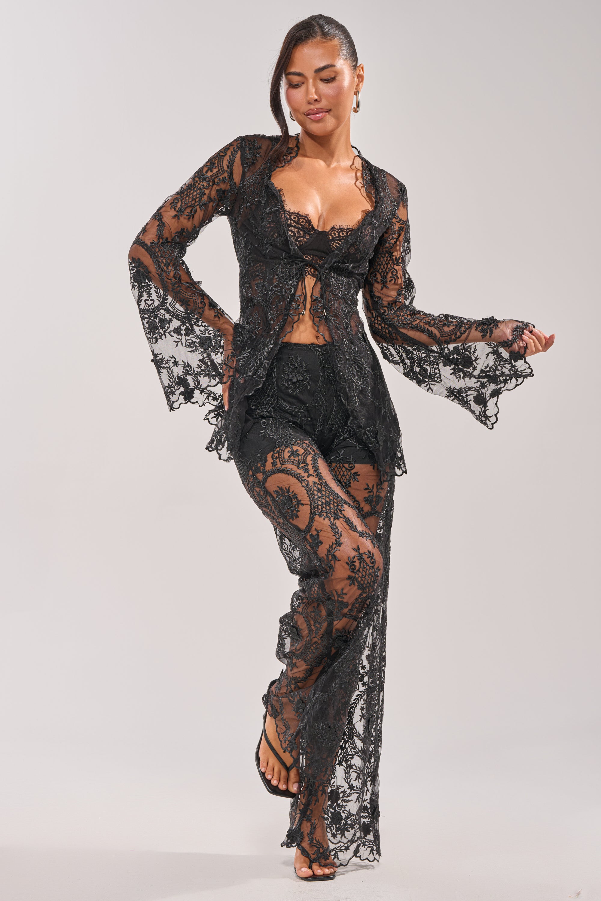 A woman poses barefoot with one foot slightly raised in the SUPER LUXE LACE TIE-FRONT BLOUSE, a black sheer lace top featuring flared sleeves, a ruffle-trimmed neckline, and intricate floral patterns, set against a light gray background. 6