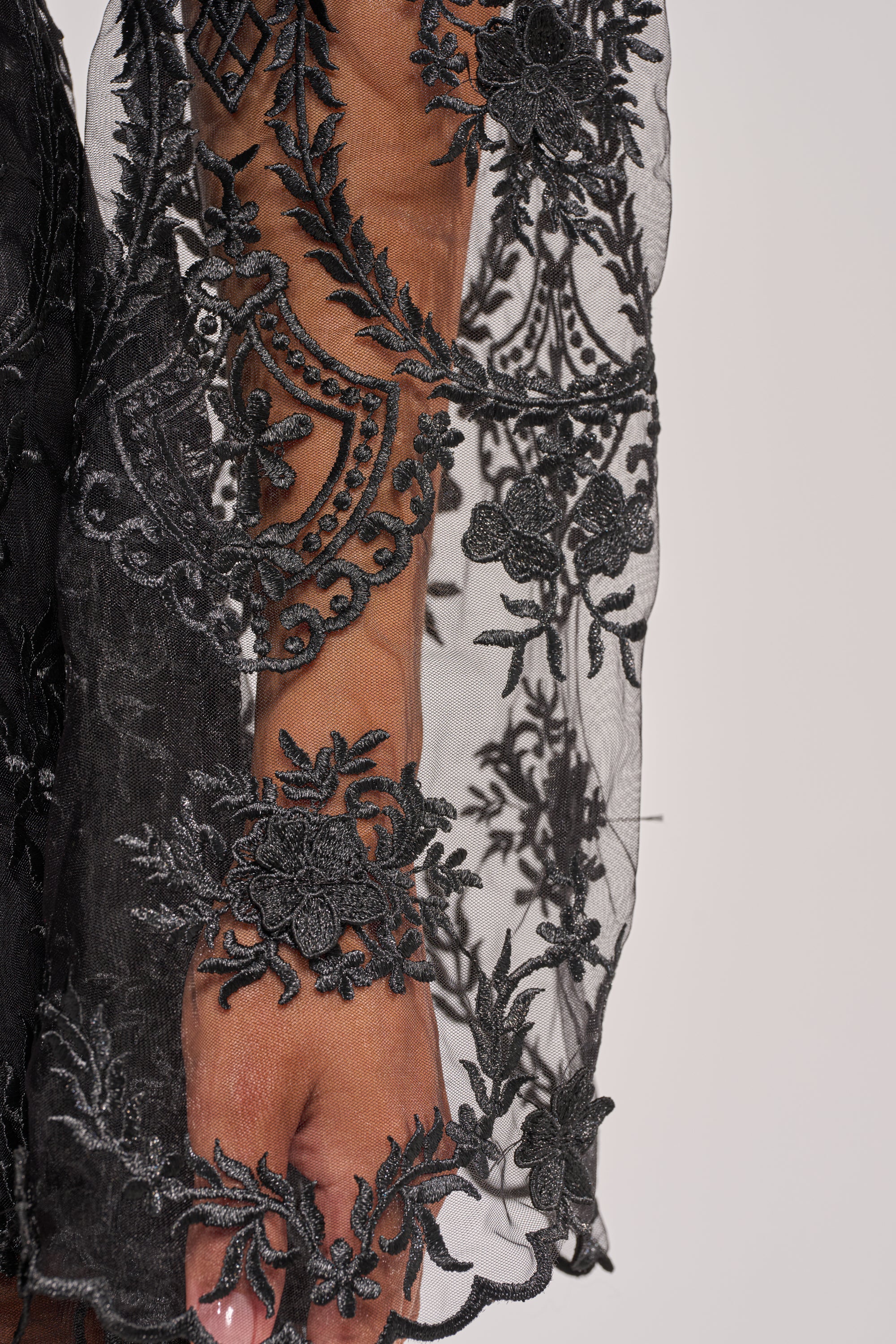 A close-up of a hand and forearm wearing the SUPER LUXE LACE TIE-FRONT BLOUSE, showcasing its black sheer lace with intricate floral and geometric embroidery. The delicate, detailed pattern is paired with manicured nails. 8