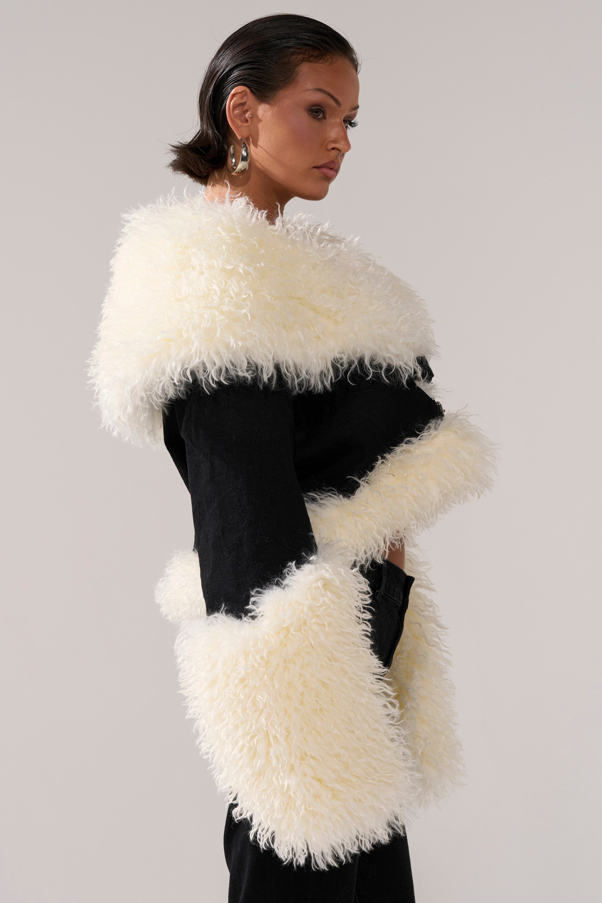 A woman with slicked-back dark hair models the SITTING PRETTY FUR TRIM DENIM JACKET, featuring a black design with oversized white faux fur on the collar, cuffs, and hem, against a plain light background.