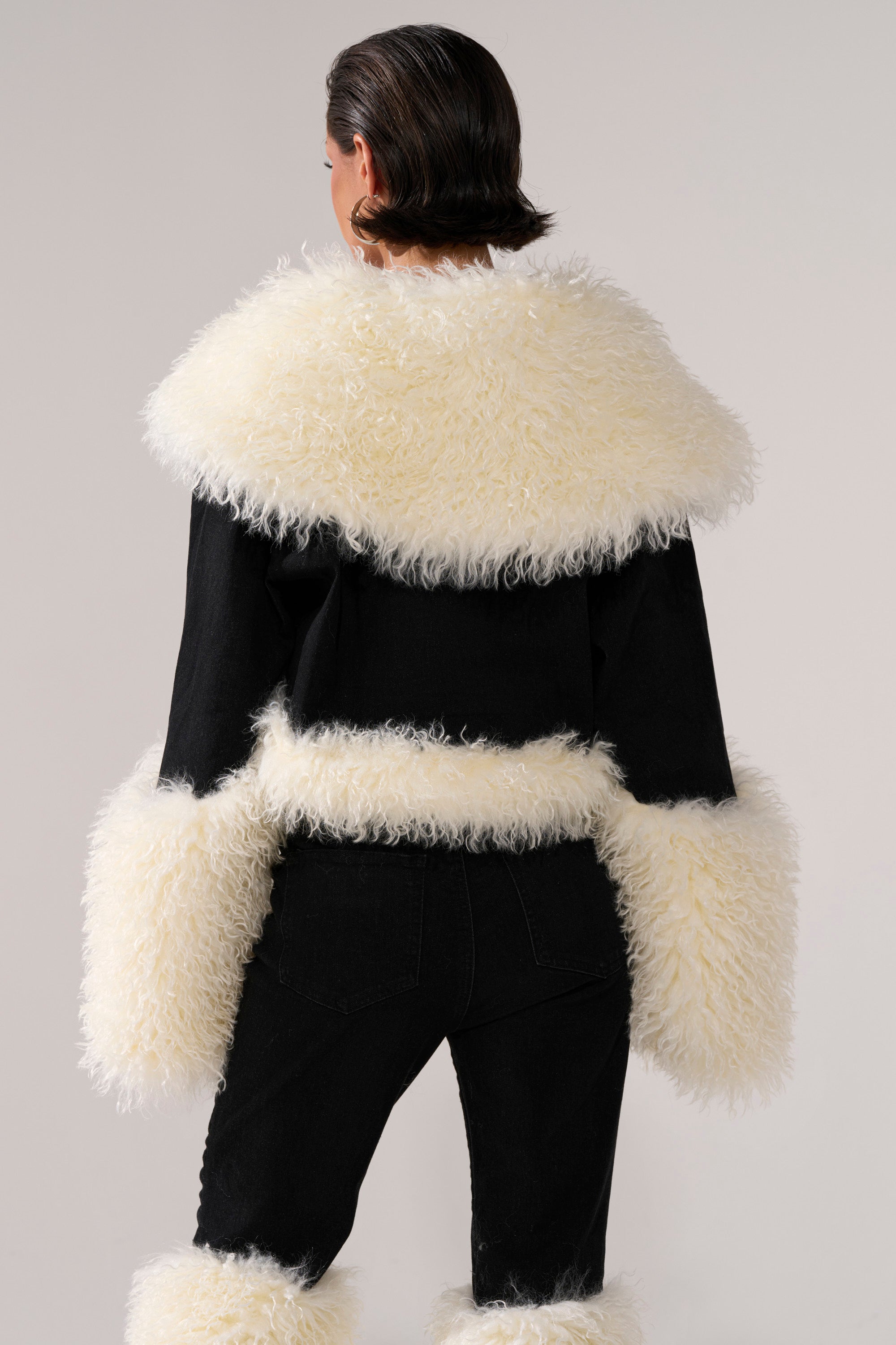 A person faces away, wearing the SITTING PRETTY FUR TRIM DENIM JACKET with large off-white faux fur on the collar and cuffs. They pair it with matching pants featuring similar trim; the background is neutral and plain.