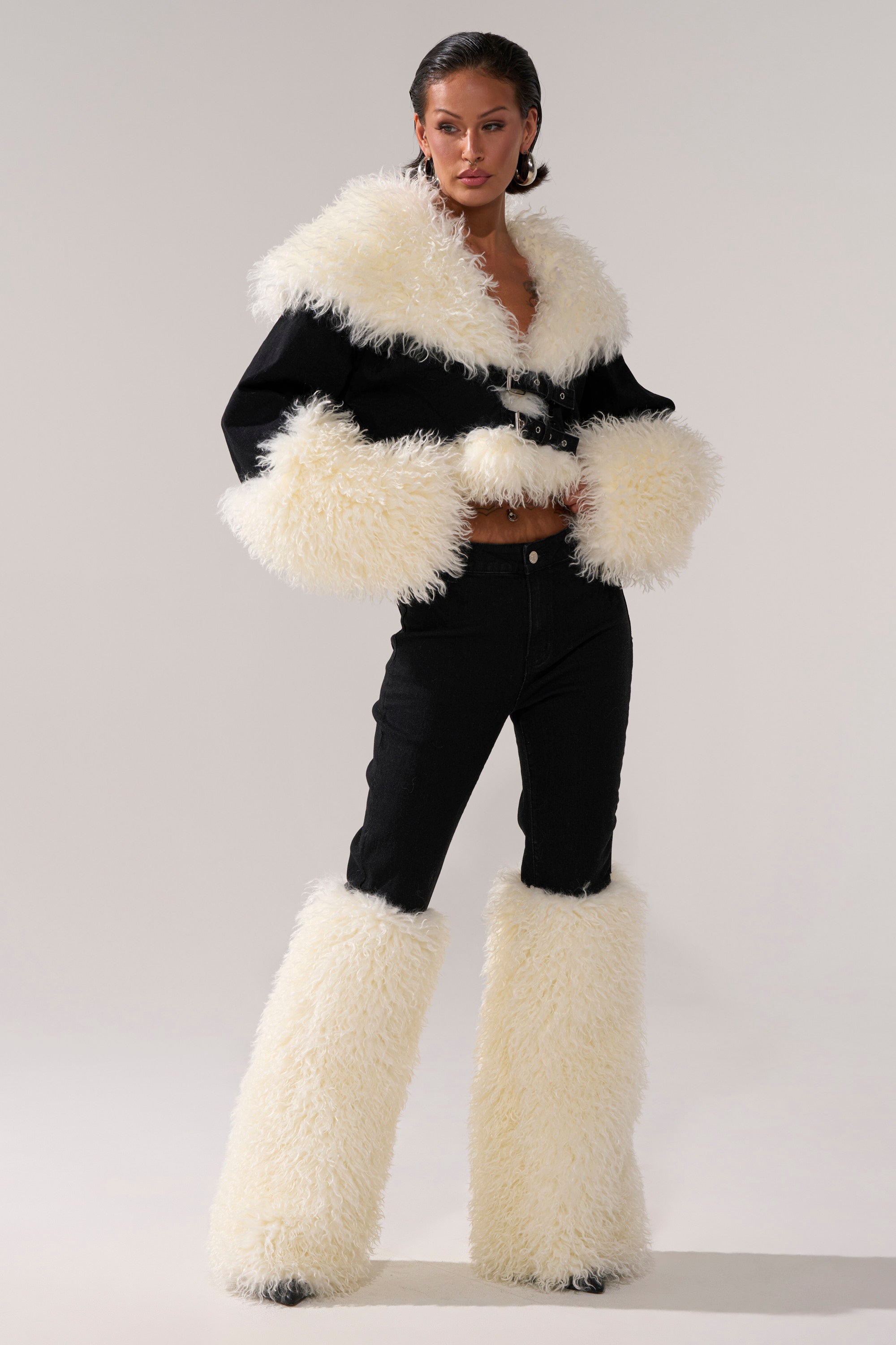 Wearing the SITTING PRETTY FUR TRIM DENIM, this person pairs a black jacket with bold white faux fur trim and matching leg warmers over black denim pants, posing confidently against a simple light background.