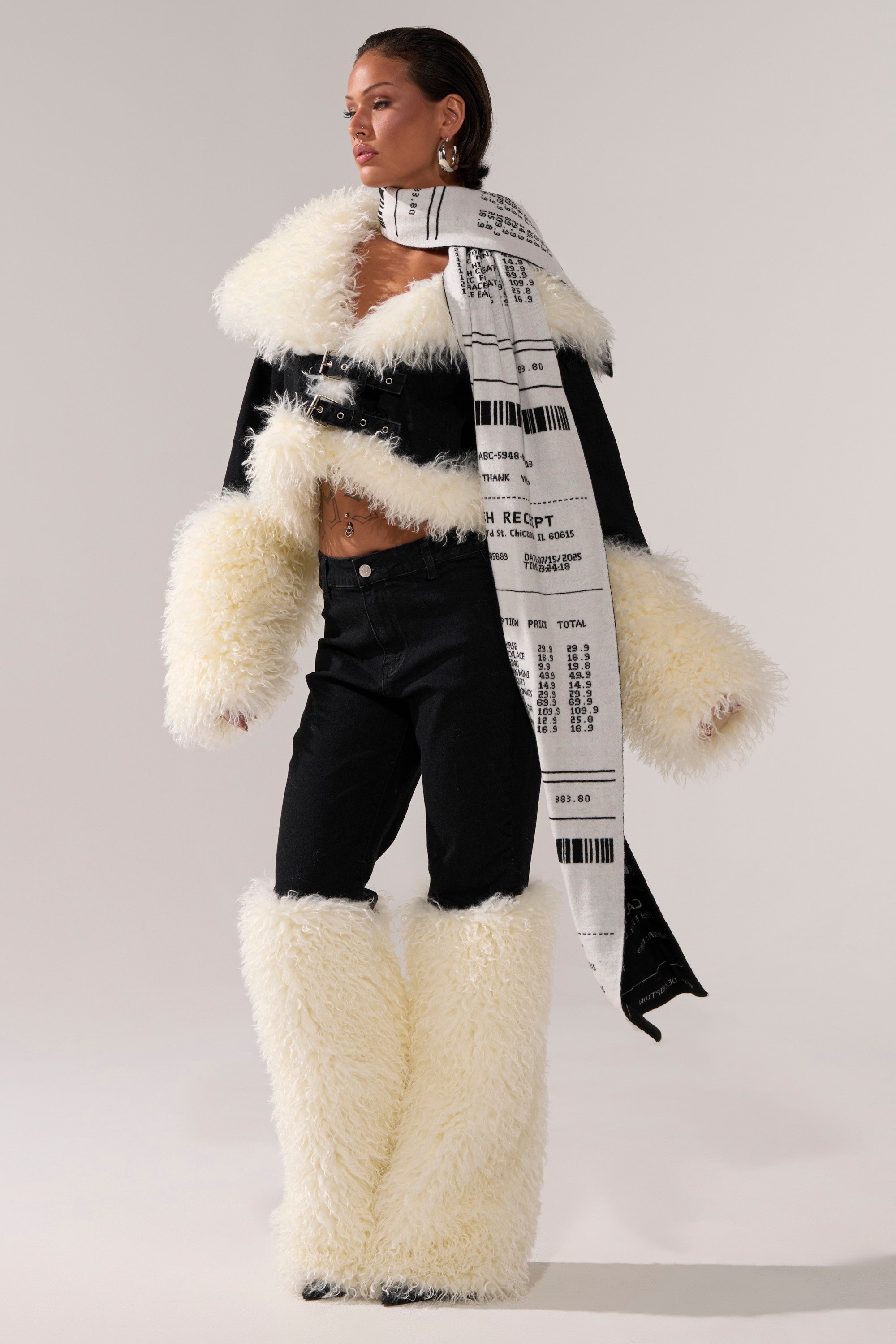 A person poses in SITTING PRETTY FUR TRIM DENIM—black high-rise jeans with large white faux fur trim—styled with a barcode receipt-print AKIRA Label scarf, plus matching jacket sleeves and boots.