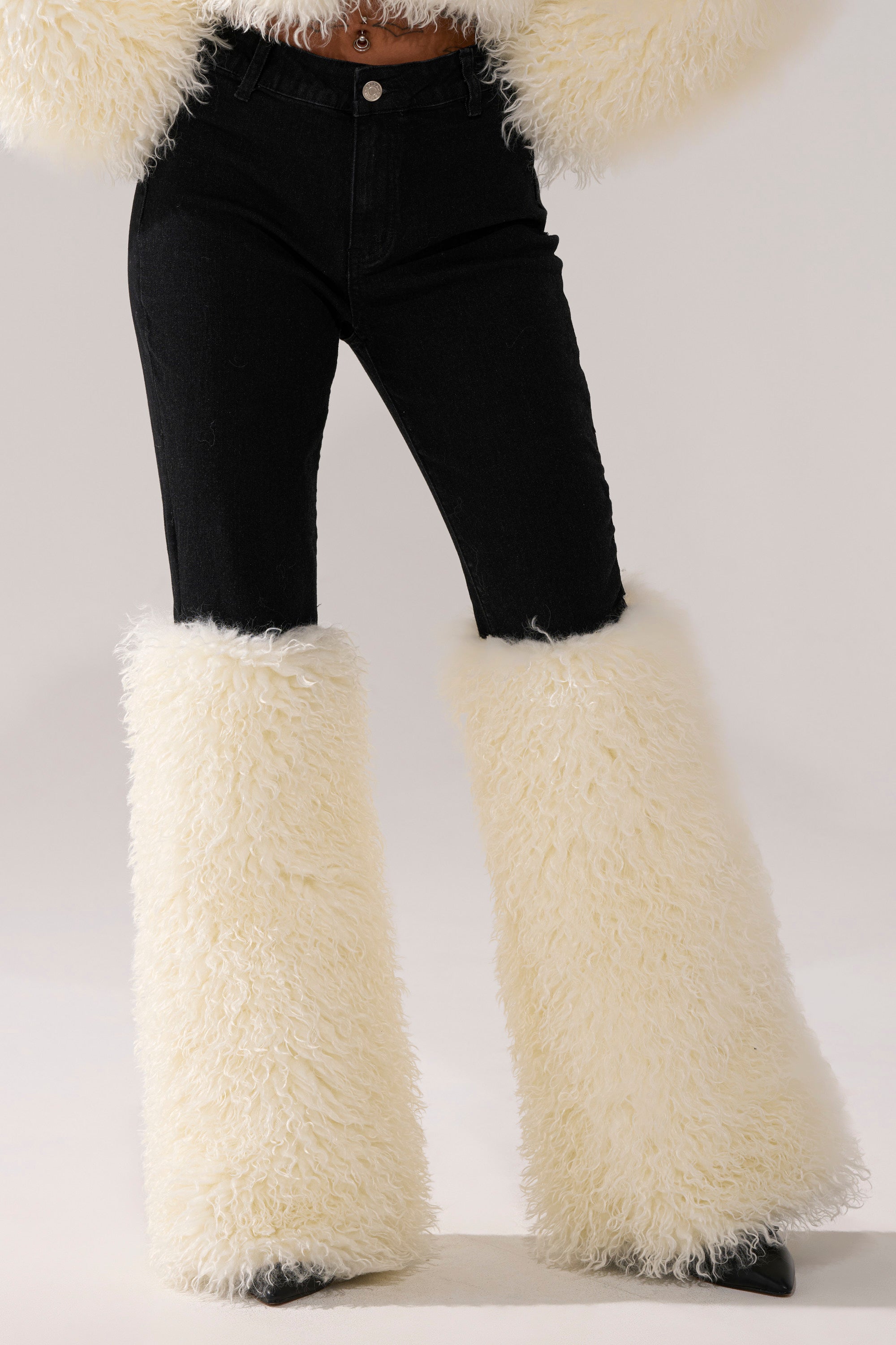 A person wears the SITTING PRETTY FUR TRIM DENIM, featuring high-rise black jeans with large, fluffy white faux fur leg warmers over boots, set against a plain light background.
