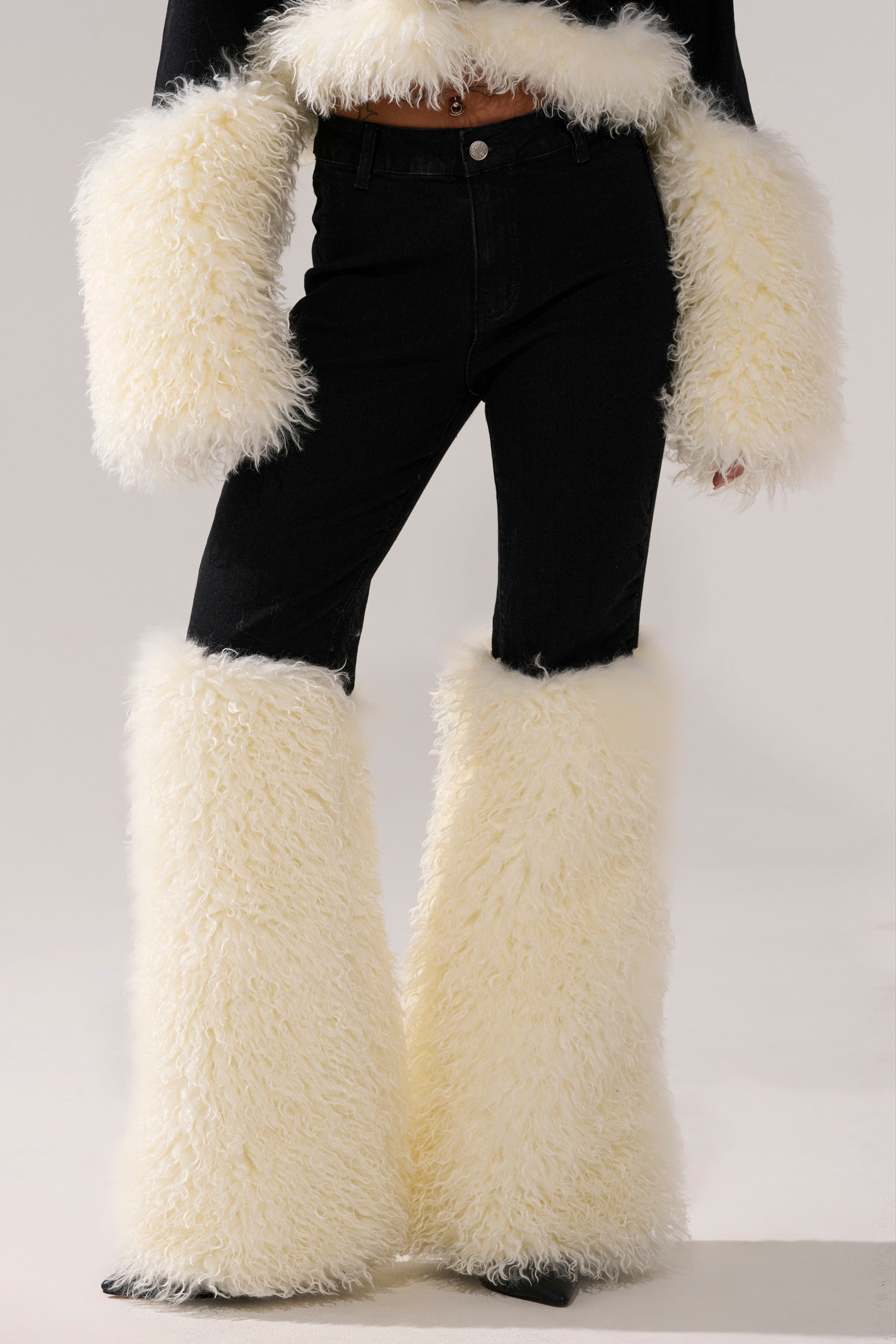 Someone wearing the SITTING PRETTY FUR TRIM DENIM: high-rise black jeans with matching cropped top, both featuring oversized cream faux fur cuffs and leg warmers, set against a neutral background.