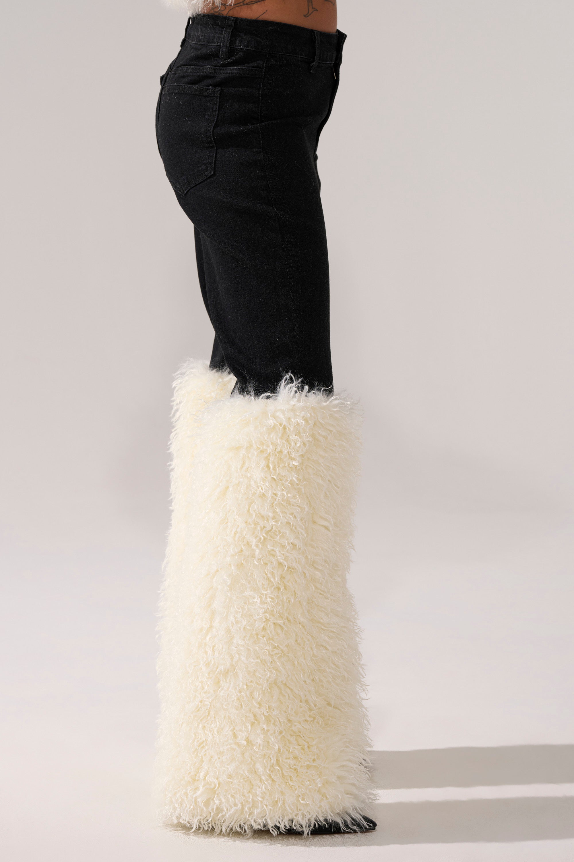 A person stands on a light background wearing SITTING PRETTY FUR TRIM DENIM—black jeans with tall, fluffy white faux fur leg warmers covering the calves.