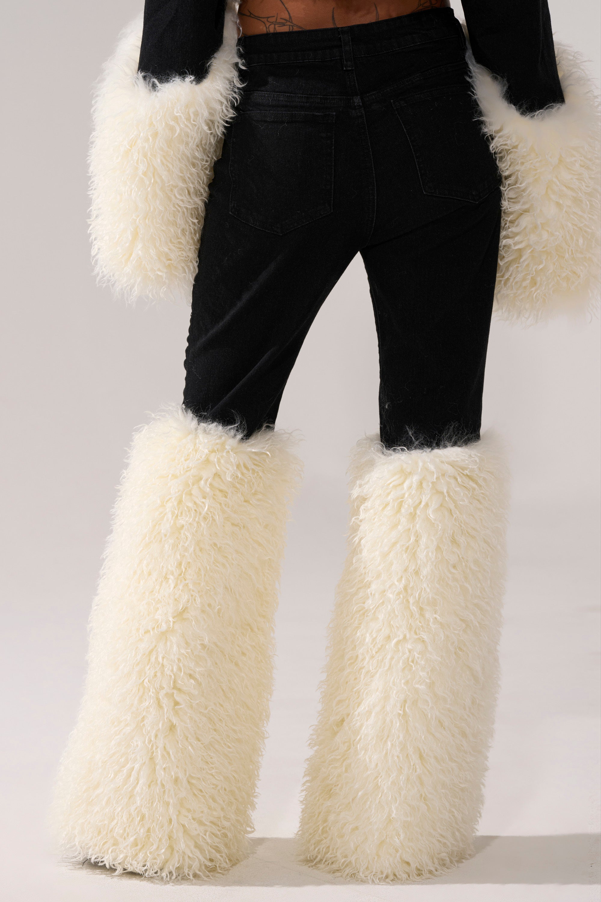 Model shown from the back wearing SITTING PRETTY FUR TRIM DENIM: black denim jeans with white fluffy faux fur leg warmers and matching fur cuffs, set against a plain background.