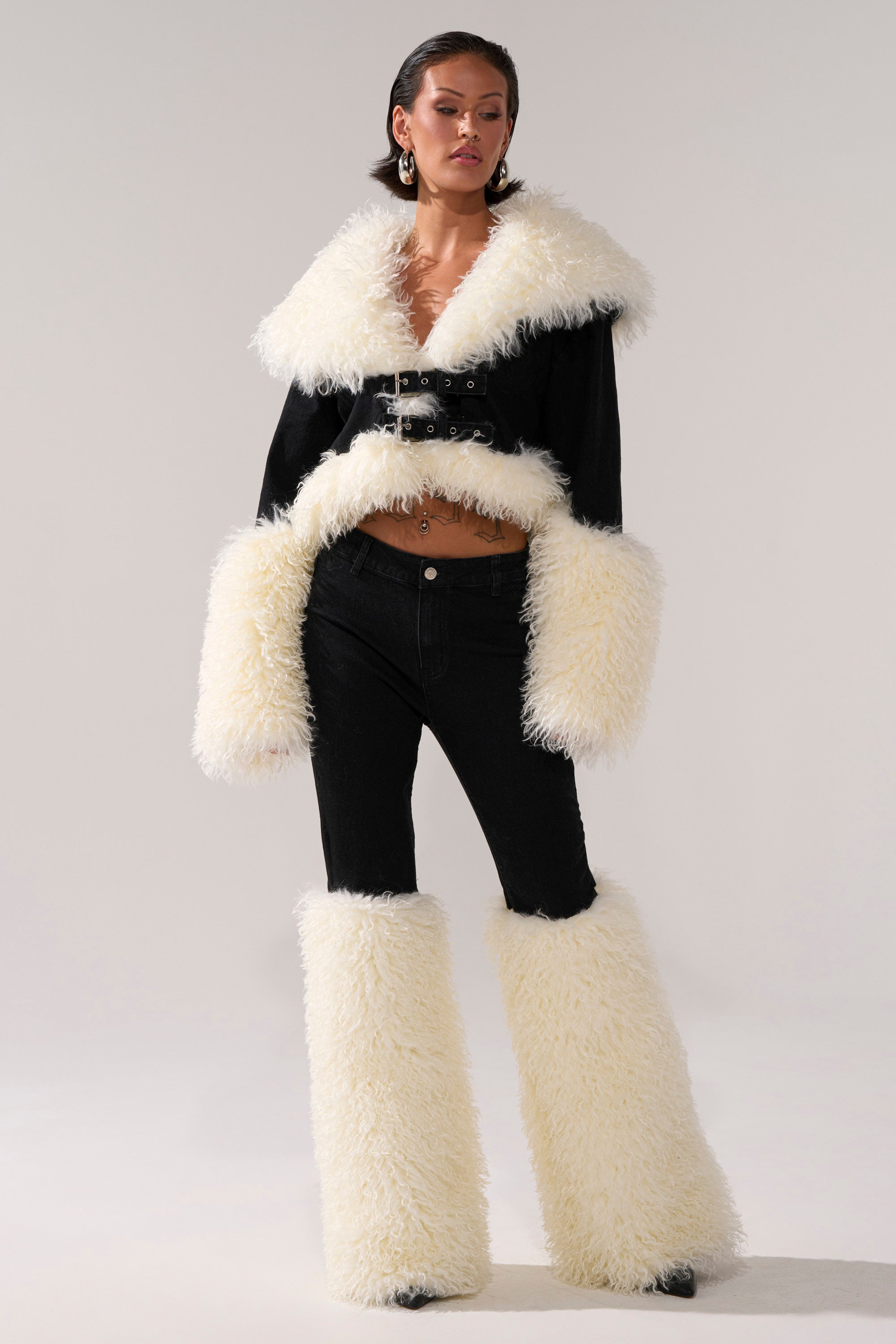 A person wears the SITTING PRETTY FUR TRIM DENIM set: a black crop top with fluffy white faux fur collar and cuffs, high-rise black jeans, and matching white faux fur leg warmers against a plain light gray background.