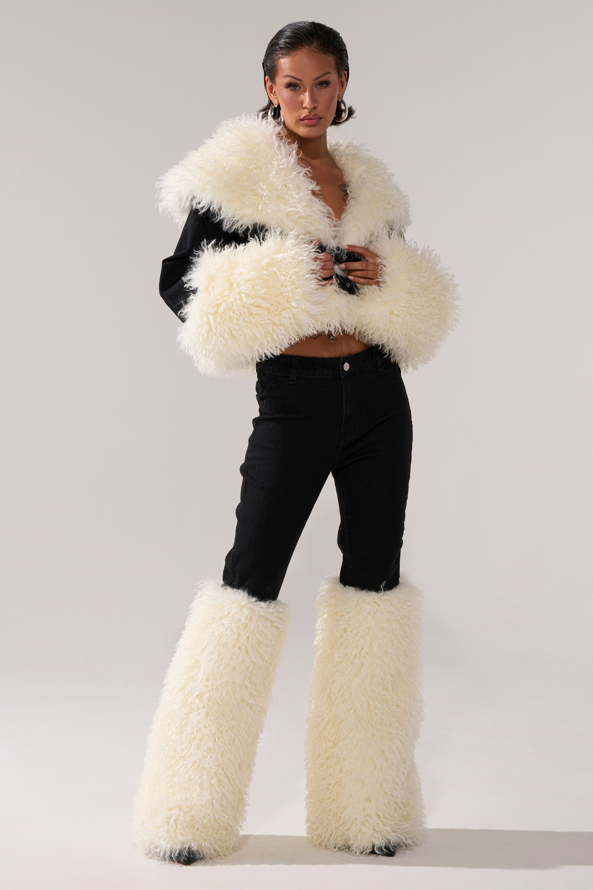A person stands confidently in the SITTING PRETTY FUR TRIM DENIM, featuring fluffy white faux fur trim leg warmers over high-rise black jeans, set against a neutral background.
