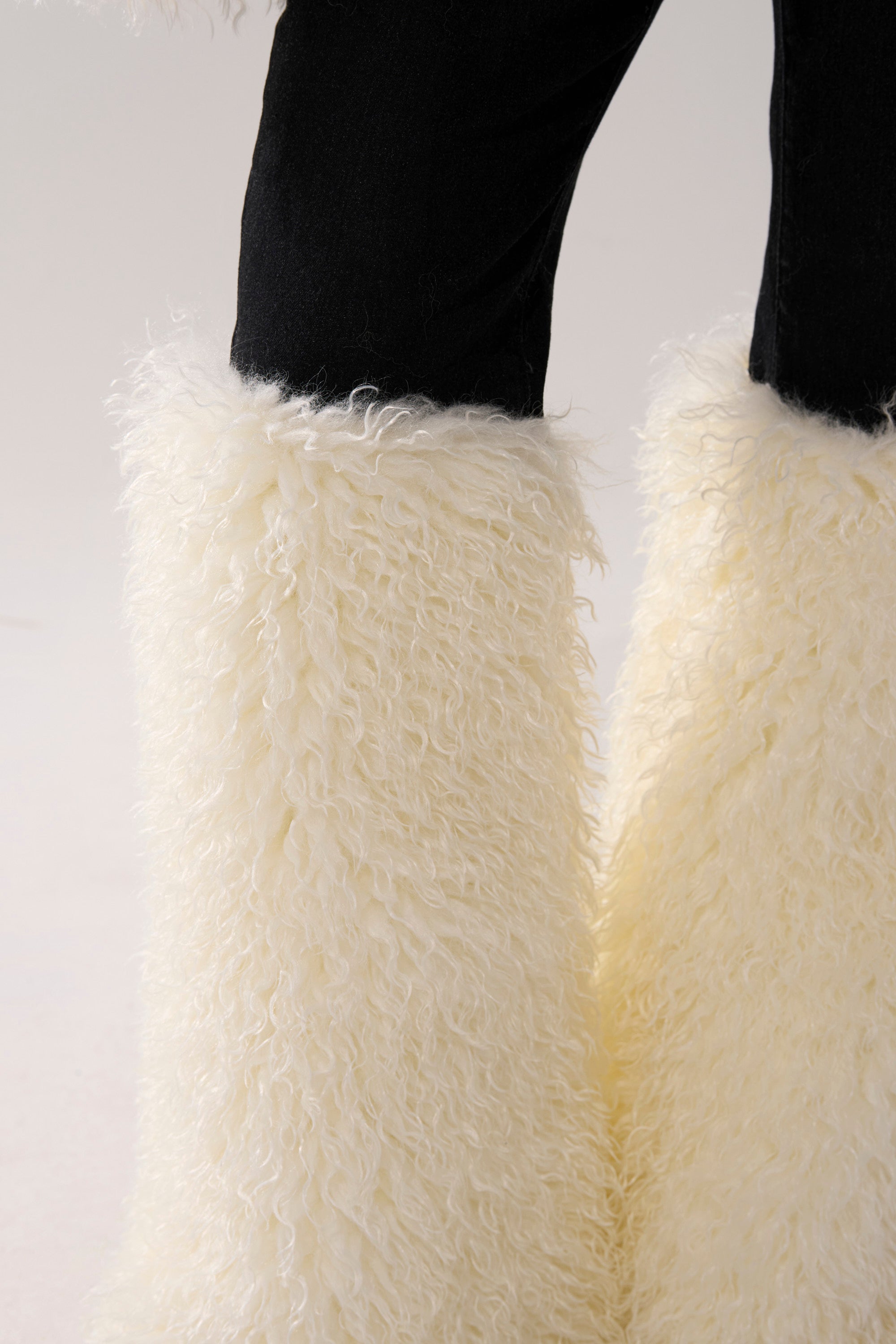 A person wears SITTING PRETTY FUR TRIM DENIM—high-rise black jeans with large white faux fur trim on the lower legs—standing against a plain background.
