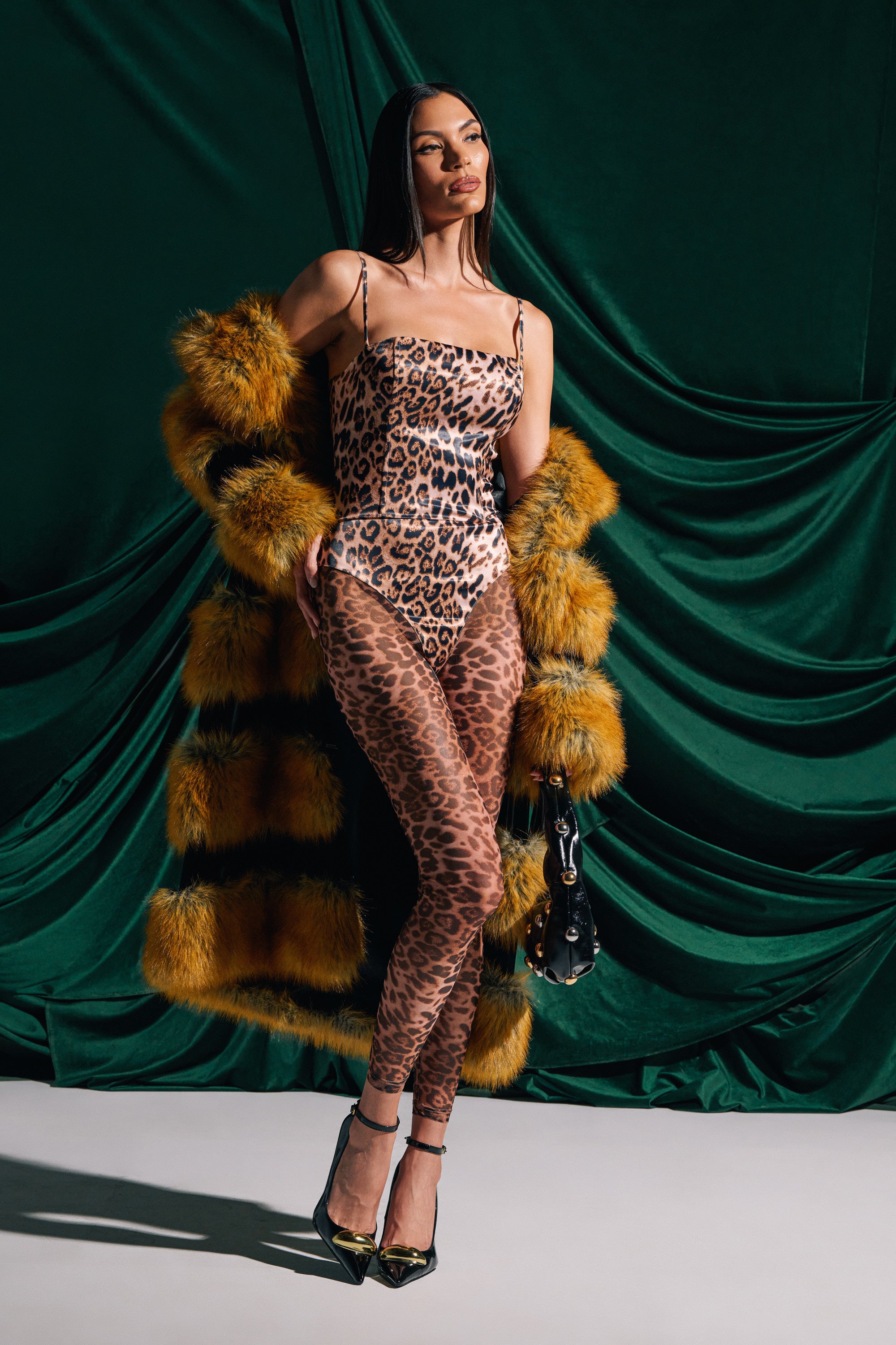 A woman poses confidently in the LENNON SATIN MESH PRINTED JUMPSUIT with black heels, a yellow faux fur coat, and a small black handbag, standing against a dark green draped fabric background.