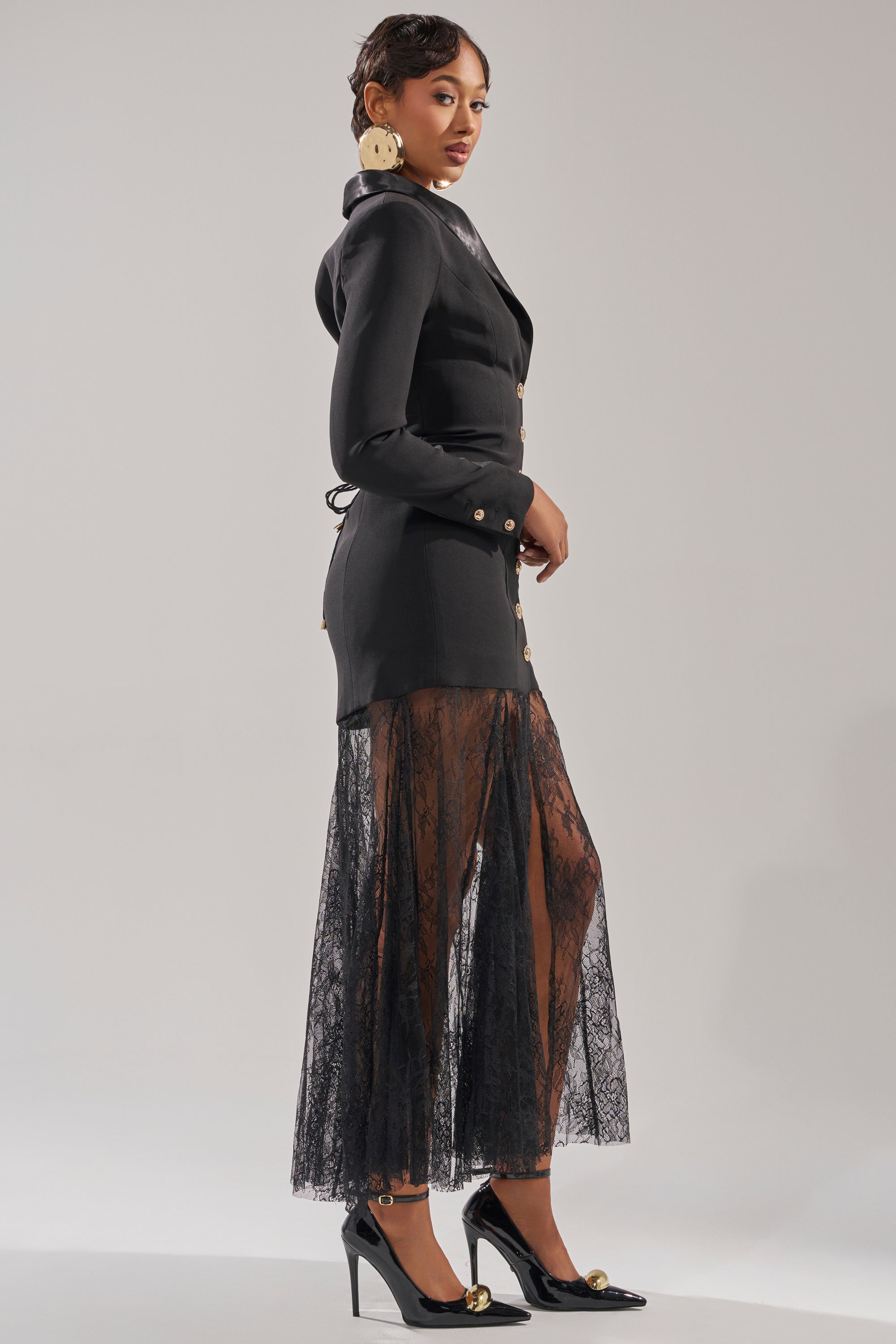 A woman wearing the OK BOSS LADY BLAZER MAXI DRESS with gold buttons and a sheer lace hem stands in profile. She pairs it with black heels featuring gold accents, large round earrings, and has her hair styled back. 3