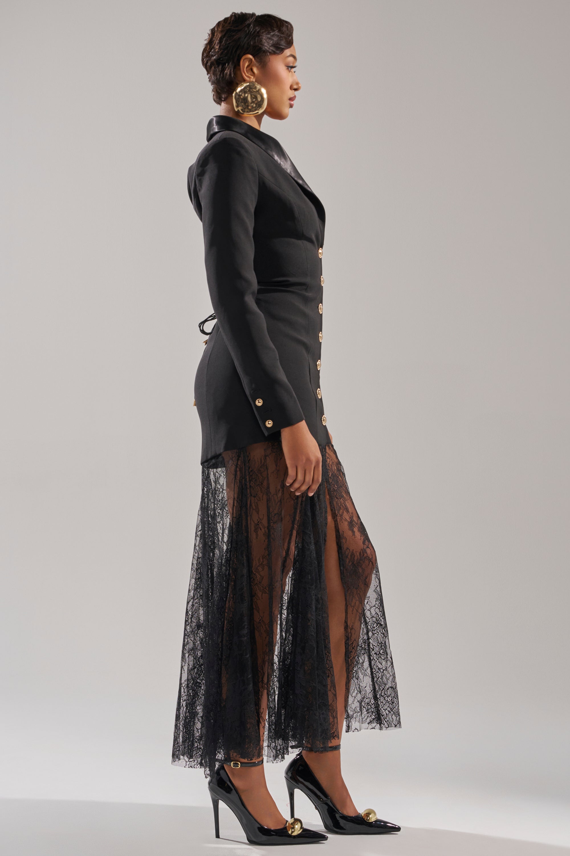 A woman in profile wears the OK BOSS LADY BLAZER MAXI DRESS featuring gold buttons and sheer lace panels from the thigh down, styled with black heels and large gold circular earrings against a neutral background. 4
