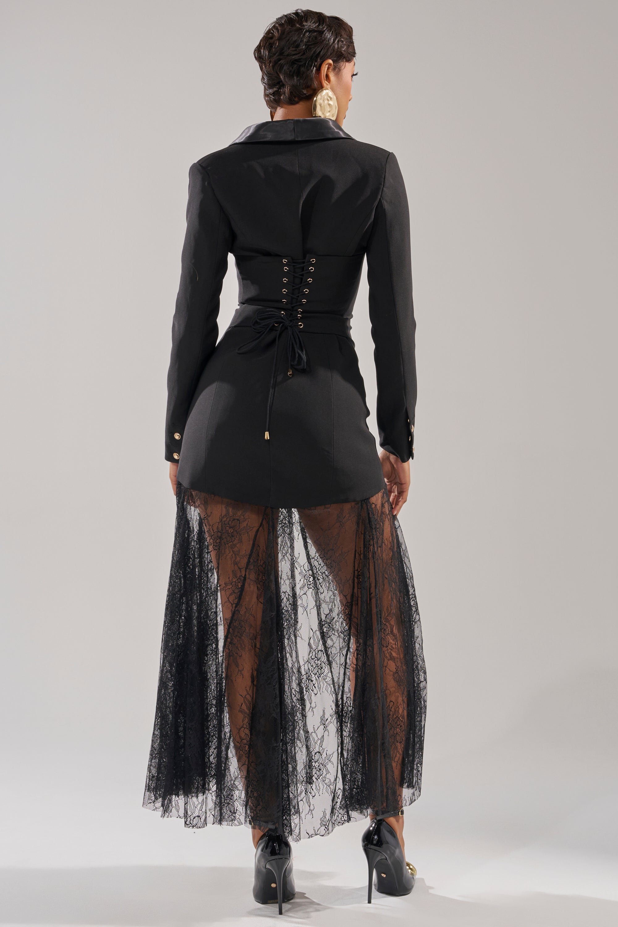 A woman faces away wearing the OK BOSS LADY BLAZER MAXI DRESS, featuring a black fitted design with lace-up details and a sheer lace skirt, styled with black high heels and bold earrings against a plain backdrop. 5
