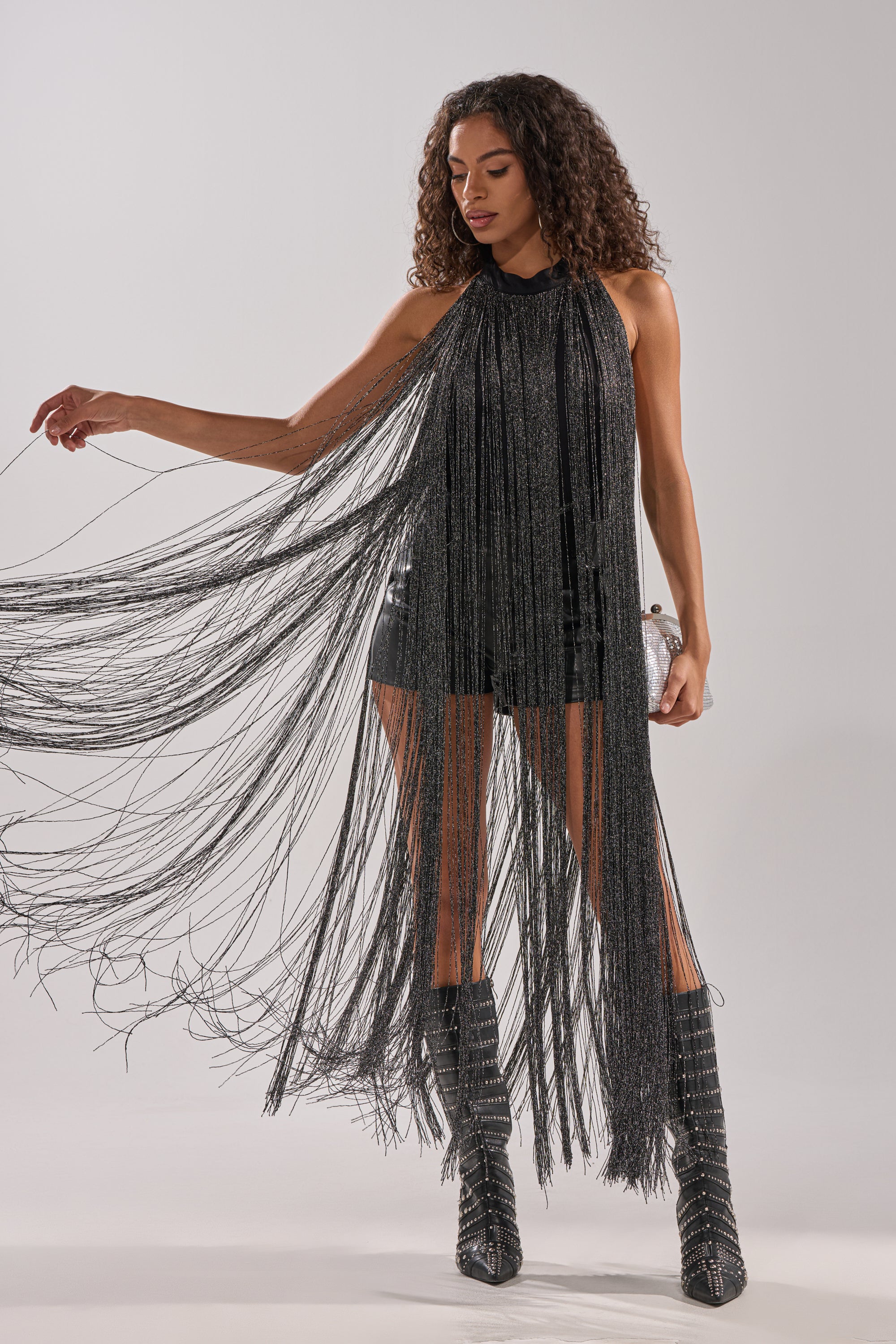A woman models the HOLIDAY FLAIR FRINGE BODYSUIT, a sleeveless black piece with long metallic fringe, paired with knee-high heeled boots. She poses gracefully, one arm extended to showcase the flowing fringe against a plain background. 0