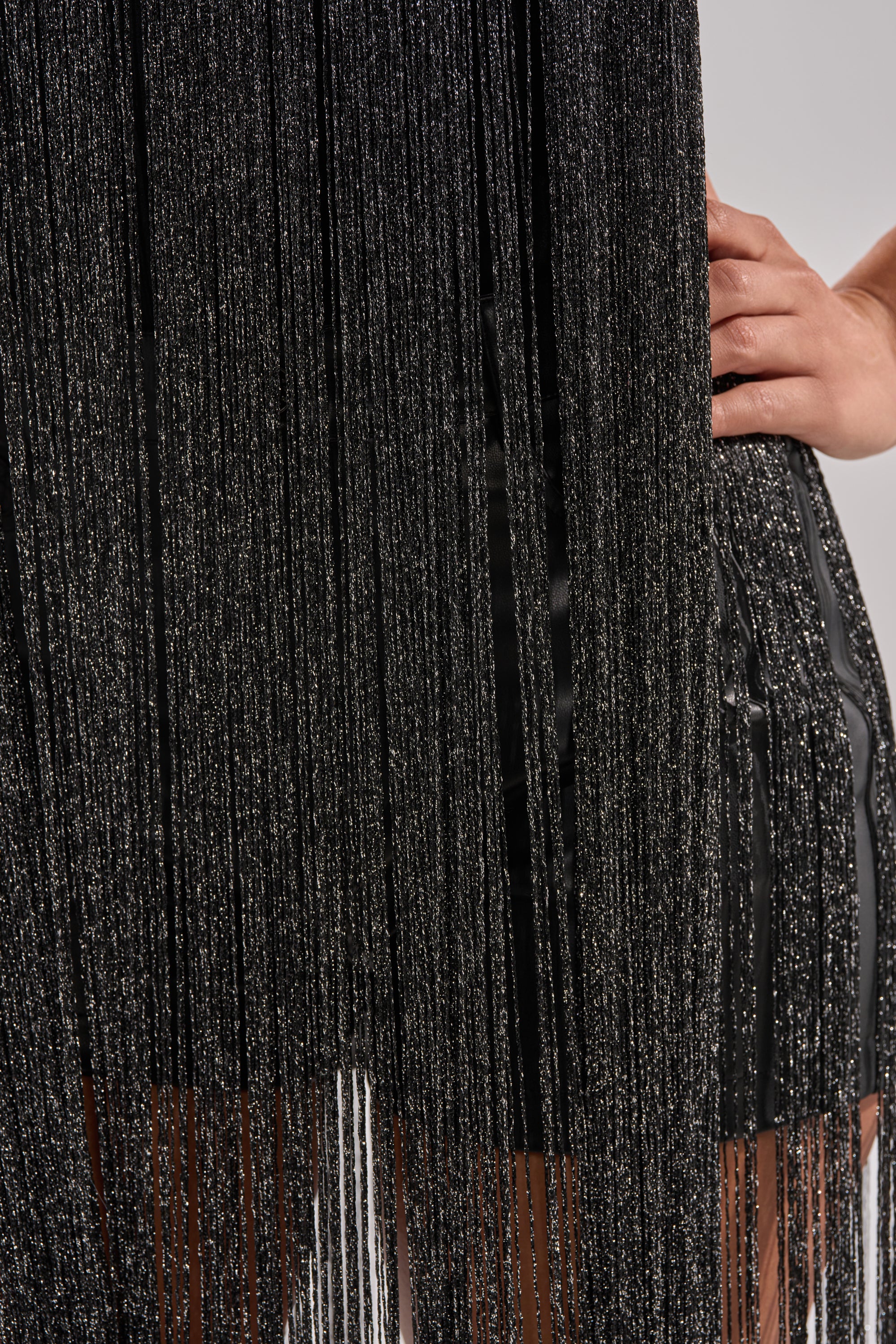 Close-up of a person wearing the HOLIDAY FLAIR FRINGE BODYSUIT in black with shimmery vertical fringe; one hand rests on their hip, set against a neutral background. 7