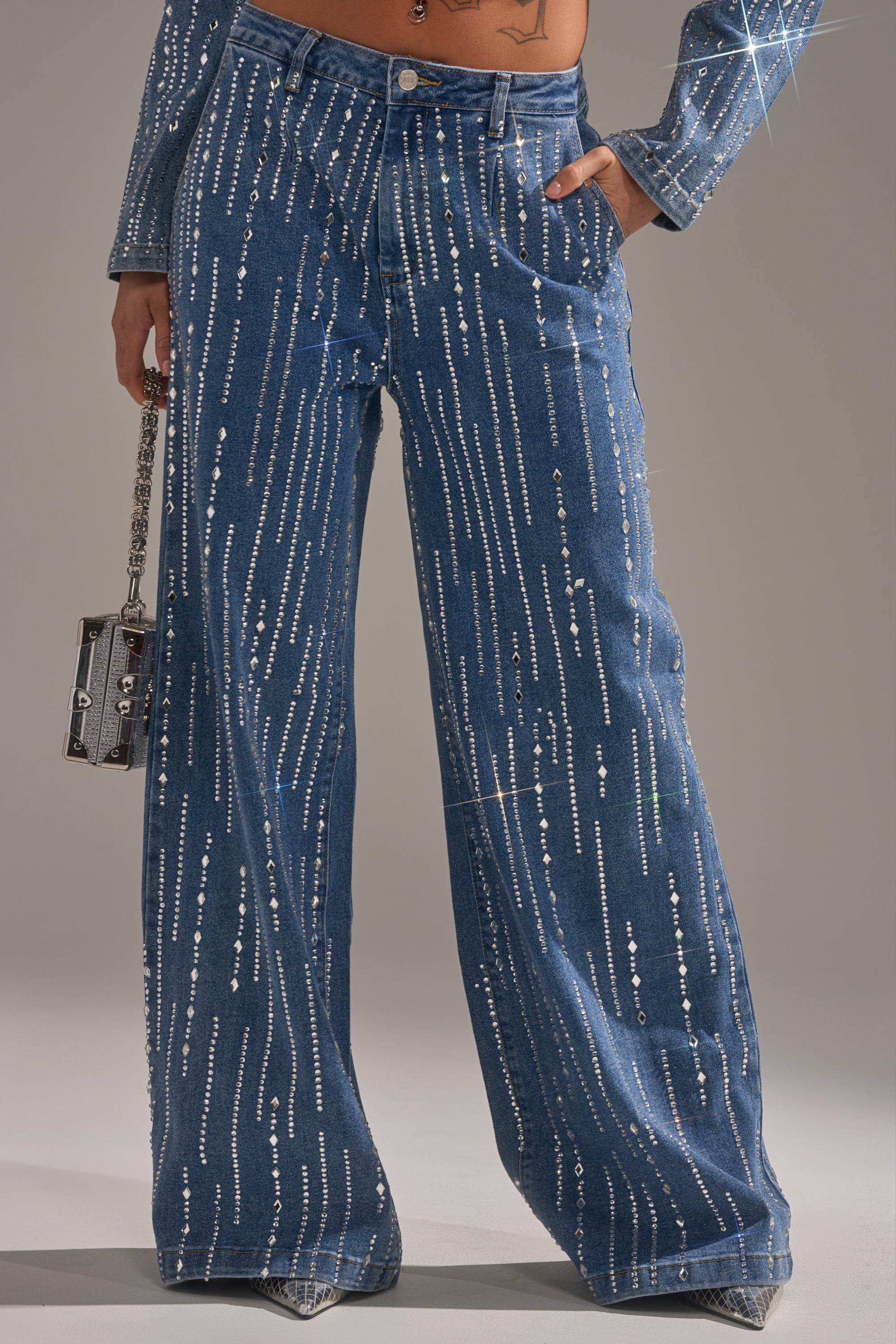 Wearing the NEVER COME DOWN RHINESTONE DENIM high-rise, wide-leg blue jeans with vertical sparkling rhinestone studs, a person holds a small handbag, revealing part of the matching jacket and a tattooed midriff.